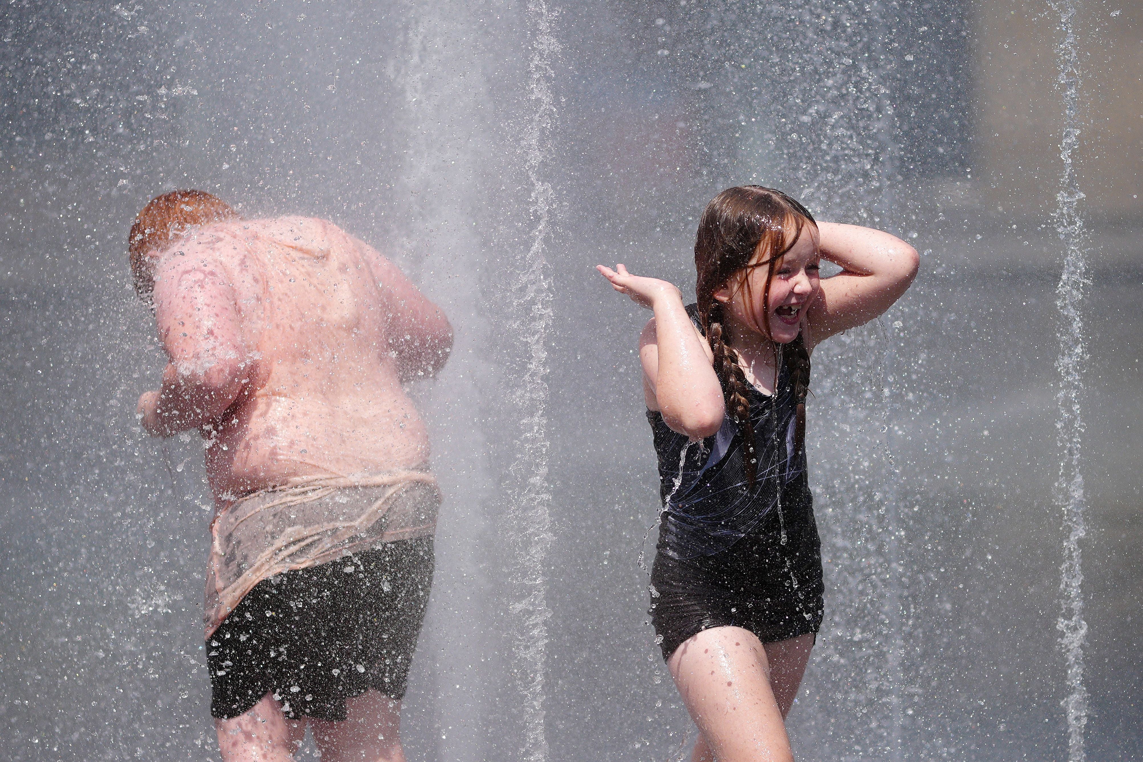 Zwei Kinder spielen lachend in den Wasserfontänen eines Parks an einem heißen Sommertag.