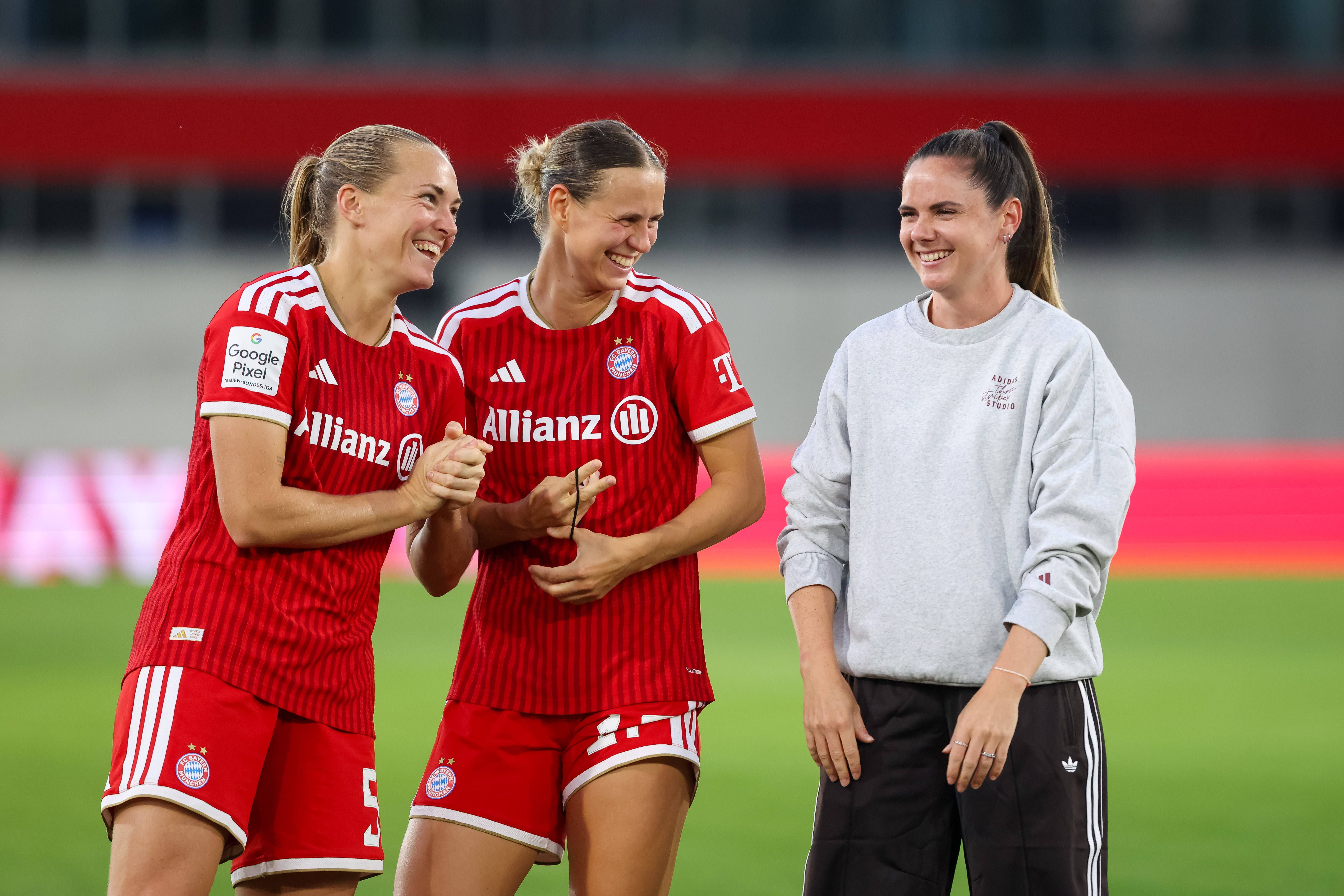 Sarah Zadrazil steht rechts im grauen Pullover lächelnd neben zwei Bayern-Teamkolleginnen im roten Trikot auf dem Spielfeld.