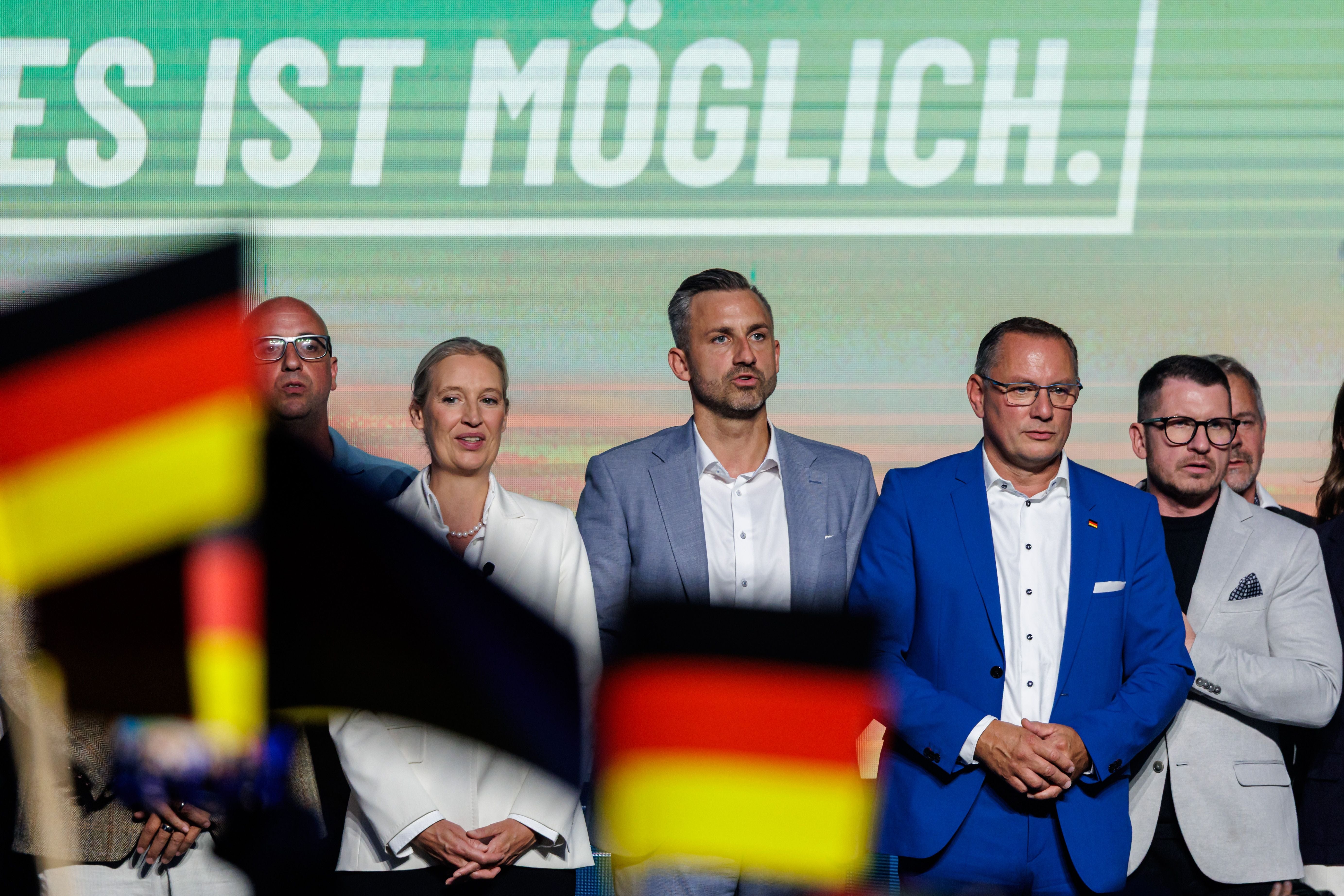 Mehrere Personen stehen auf einer Bühne, deutsche Fahnen sind im Vordergrund sichtbar.