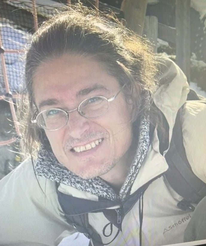 Ein Mann mit Brille, langen Haaren und Outdoorjacke lächelt in die Kamera.