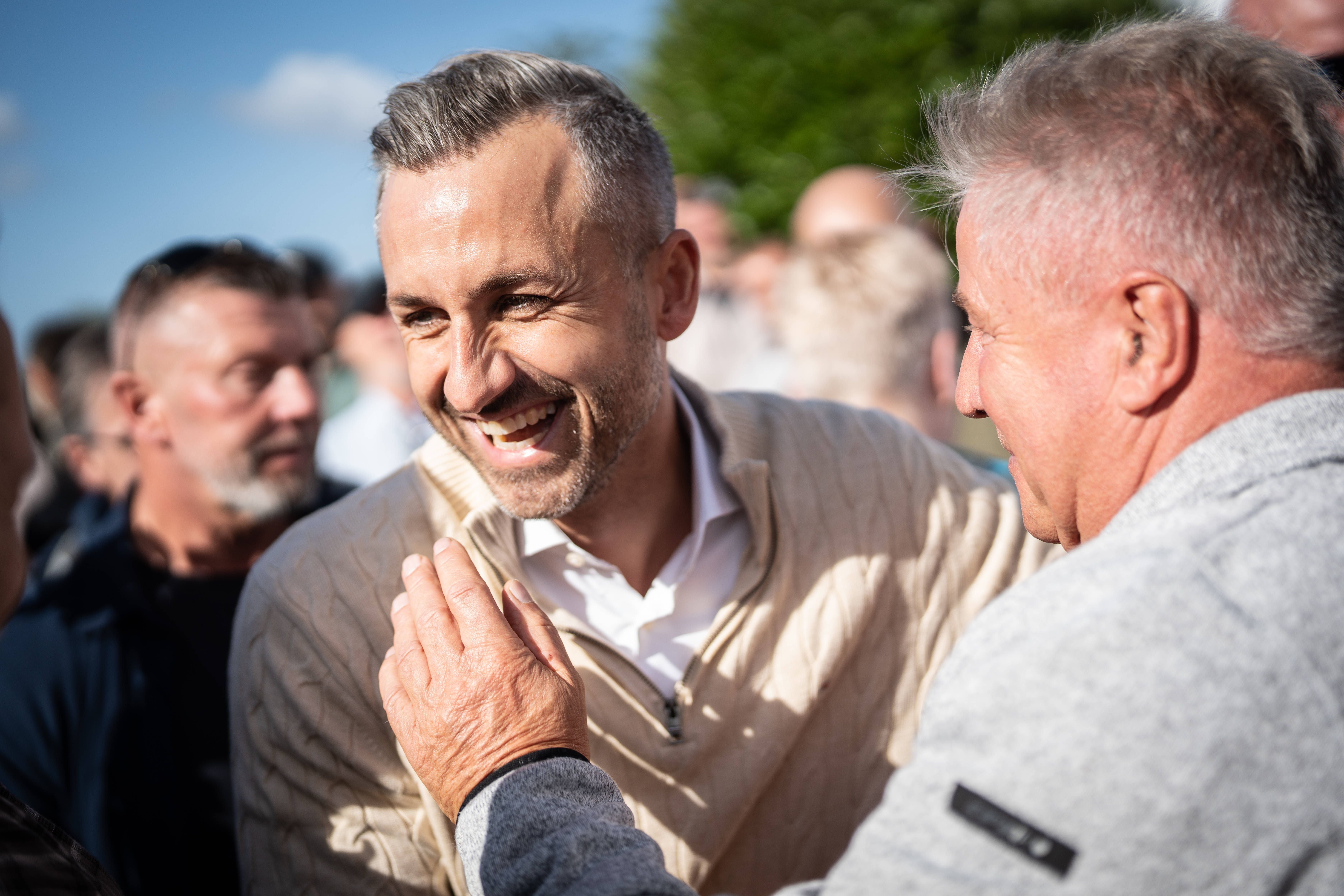 Ein lachender Mann wird im Freien herzlich von einem anderen Mann begrüßt, umgeben von einer Menschenmenge.