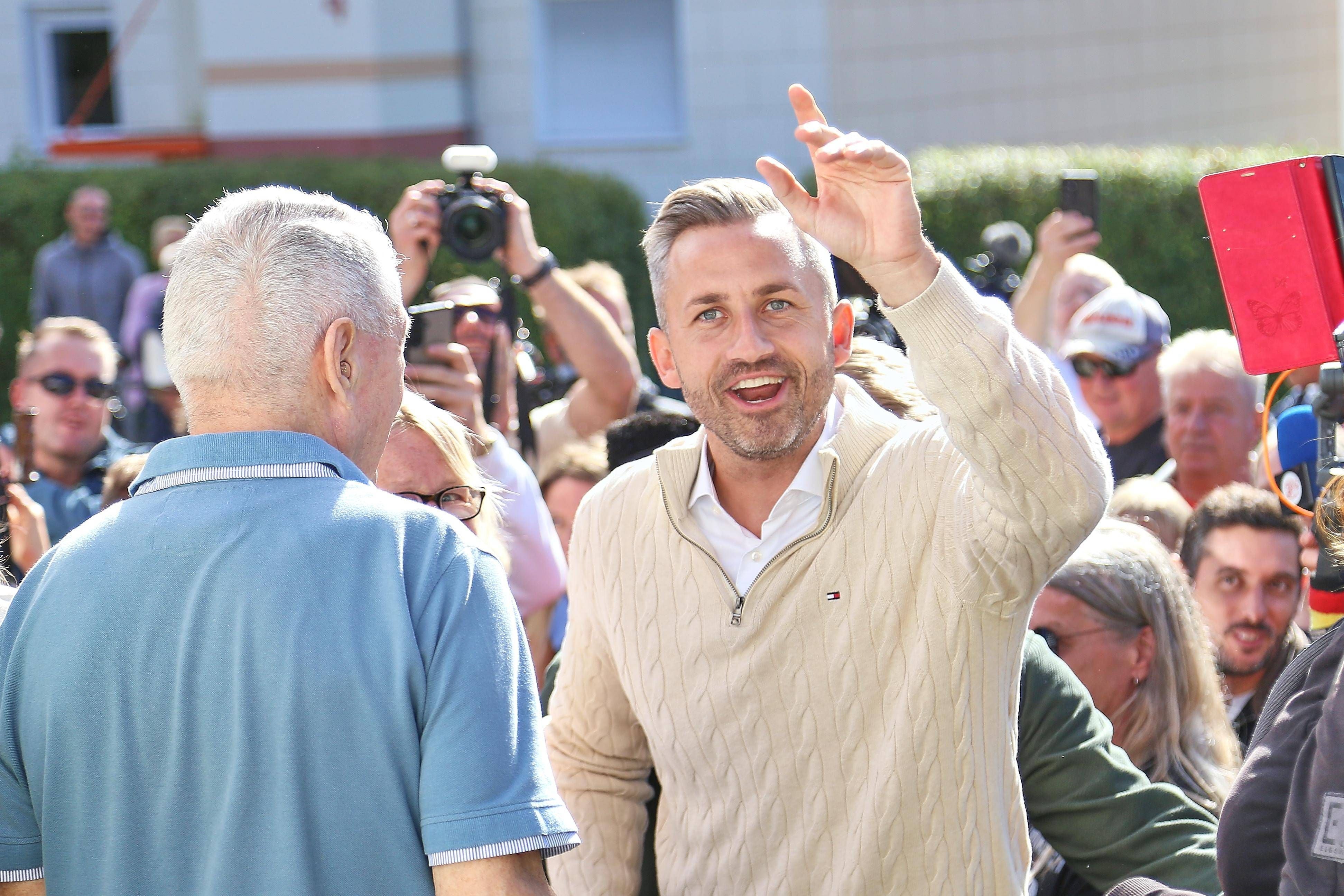 Ein Mann winkt in die Kamera, umgeben von einer Menschenmenge bei einer Wahlveranstaltung.
