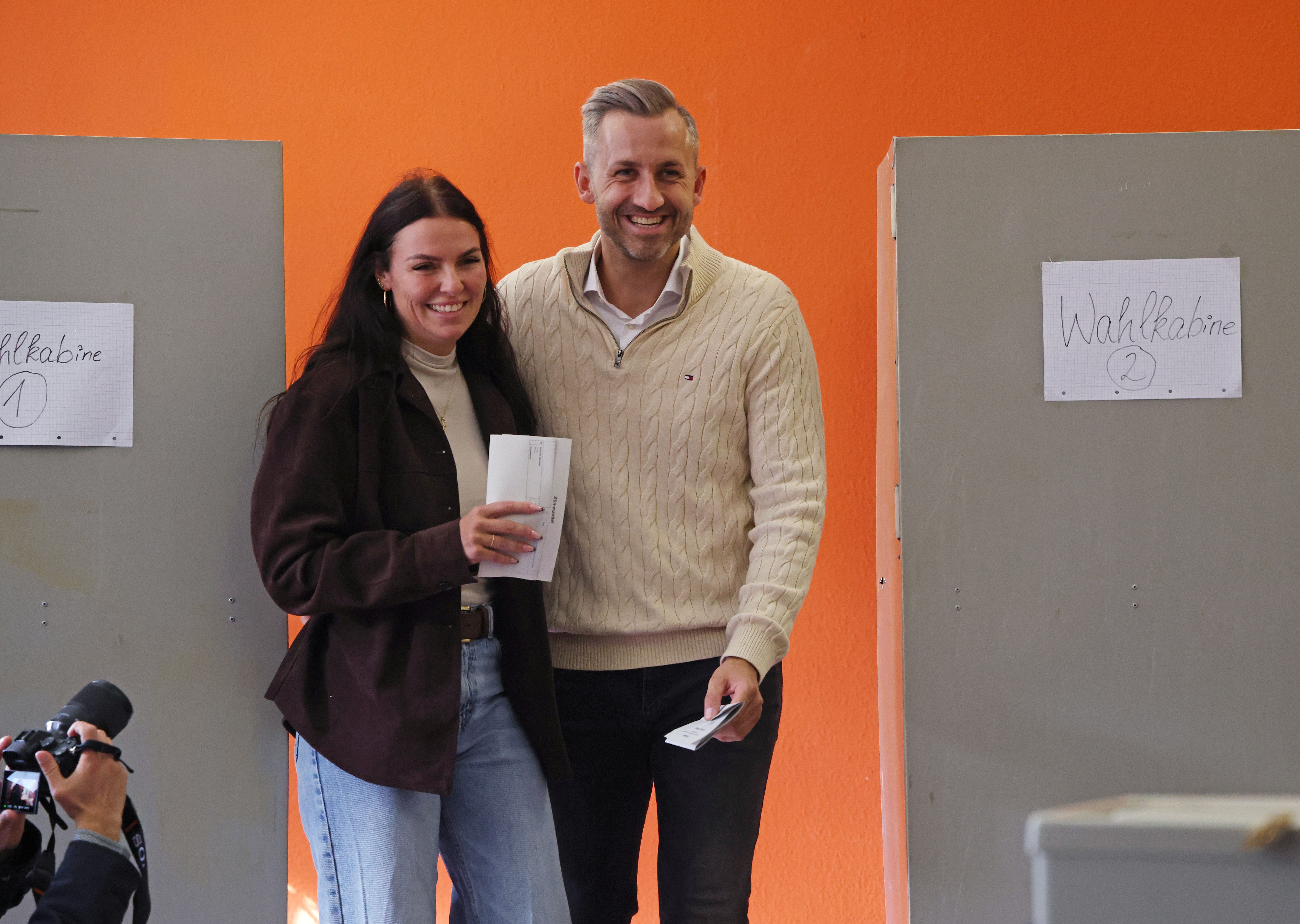 Ein lächelndes Paar wirft Wahlzettel an Wahlkabinen mit orangener Wand im Hintergrund ein.
