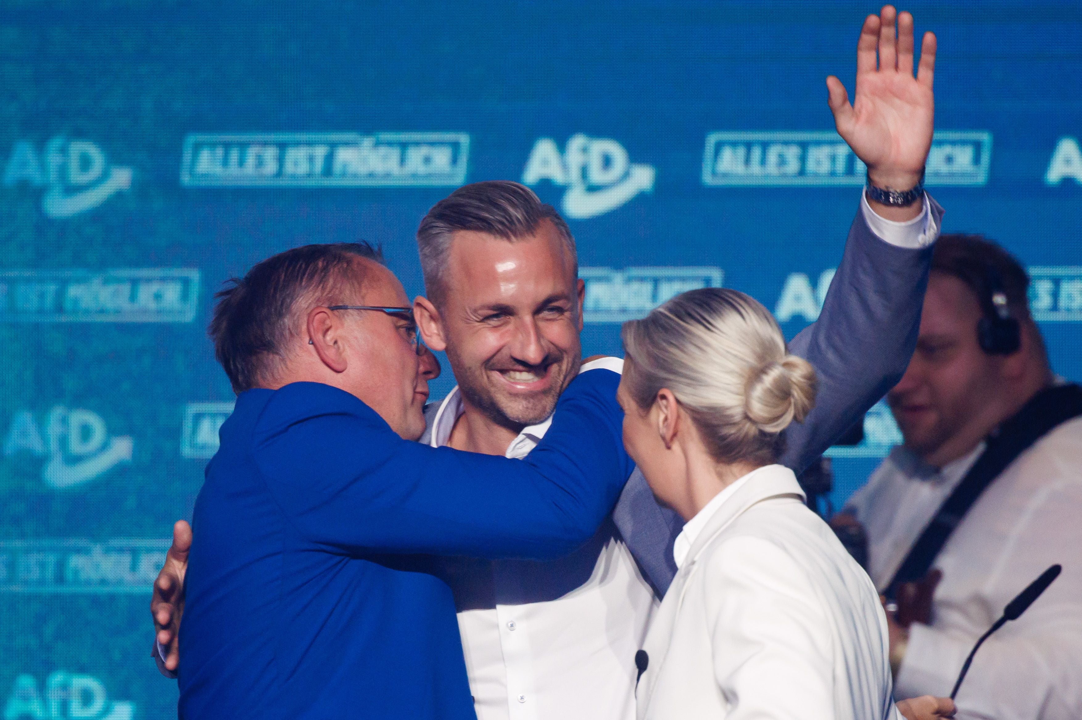 Drei Menschen umarmen sich auf einer Bühne mit AfD-Logo im Hintergrund; einer winkt lächelnd.