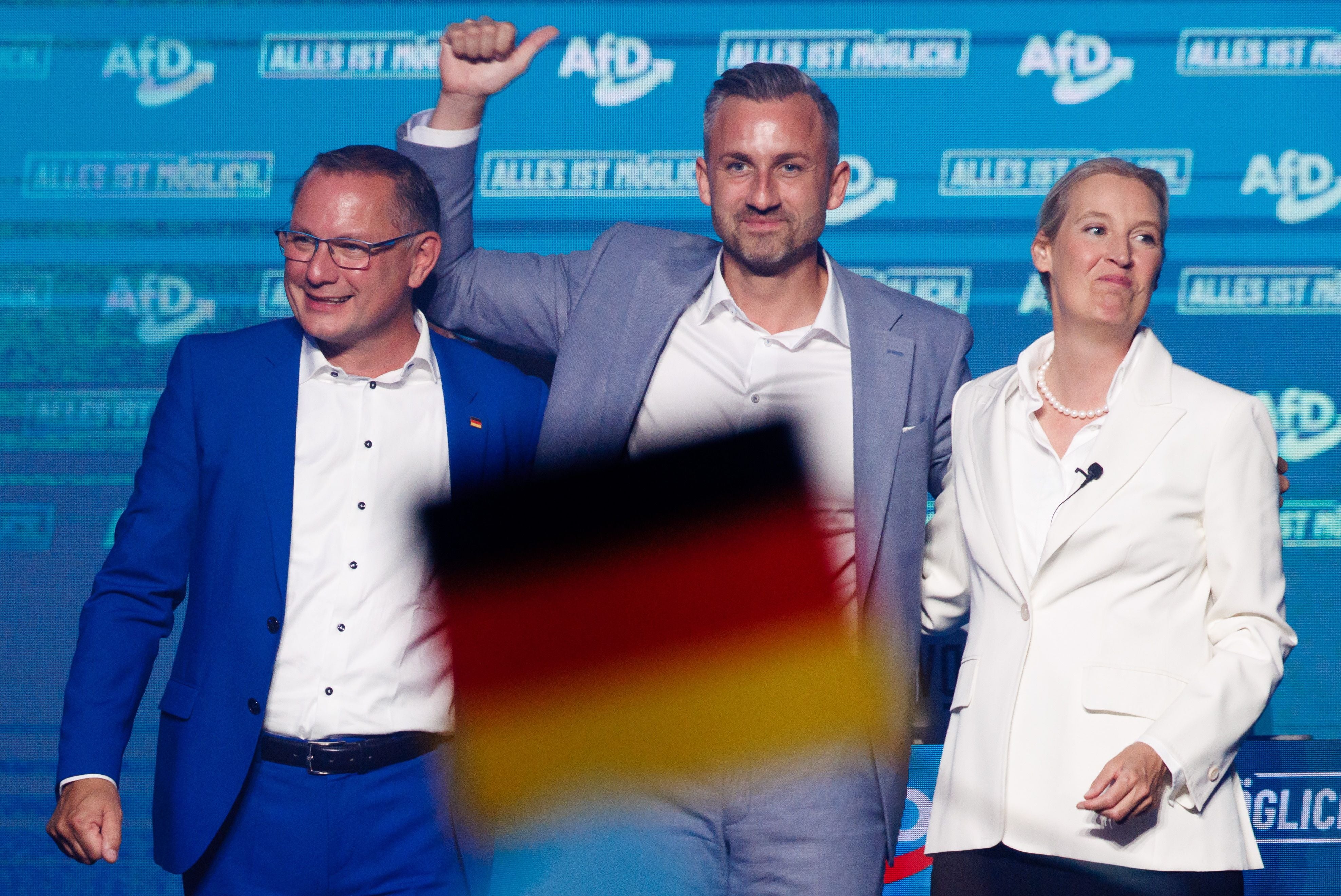 Drei Personen auf einer AfD-Bühne, im Vordergrund schwenkt jemand eine Deutschlandflagge.