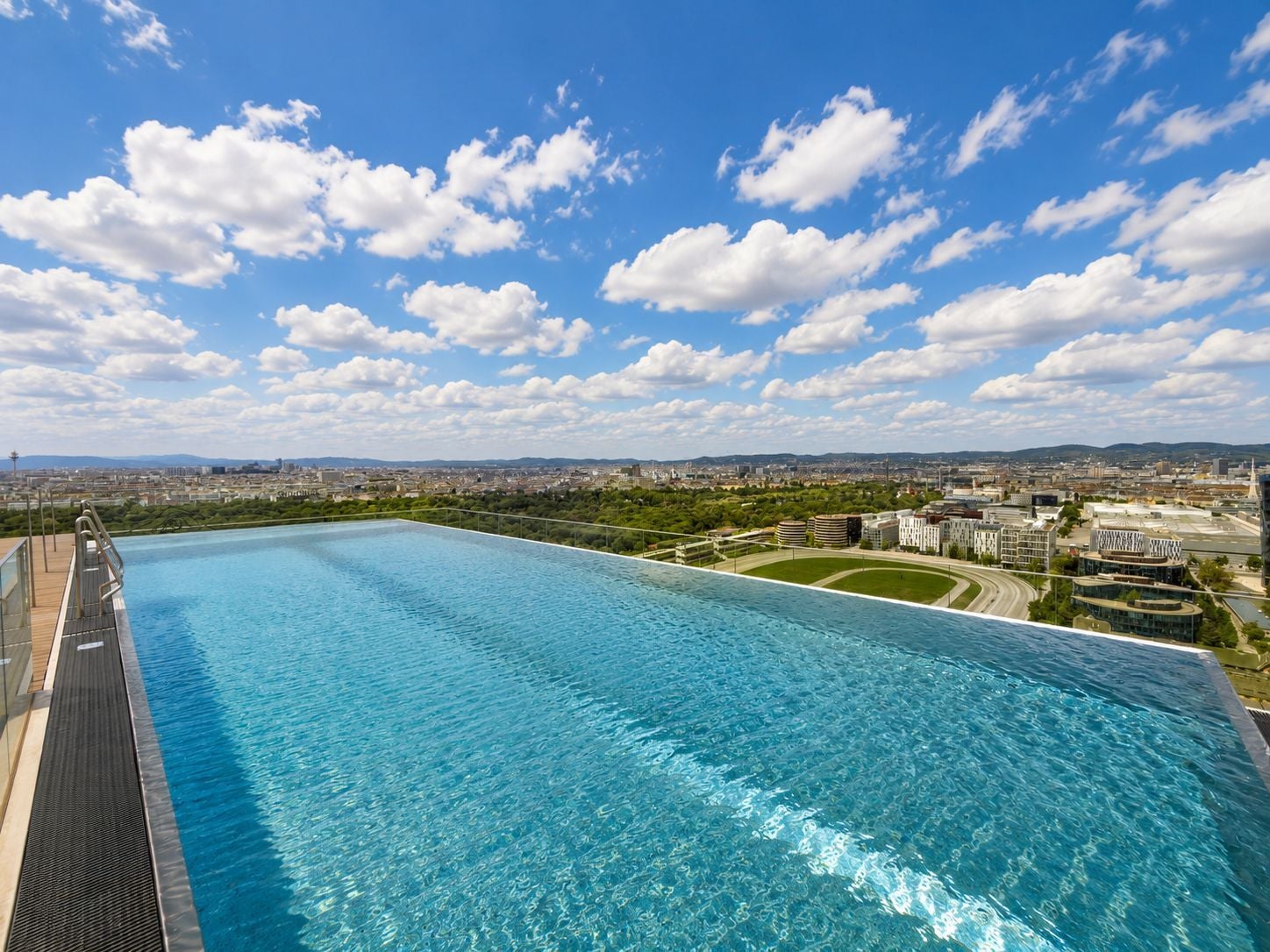Infinity-Pool auf einem Dach mit Blick über eine Stadt bei blauem Himmel und weißen Wolken.