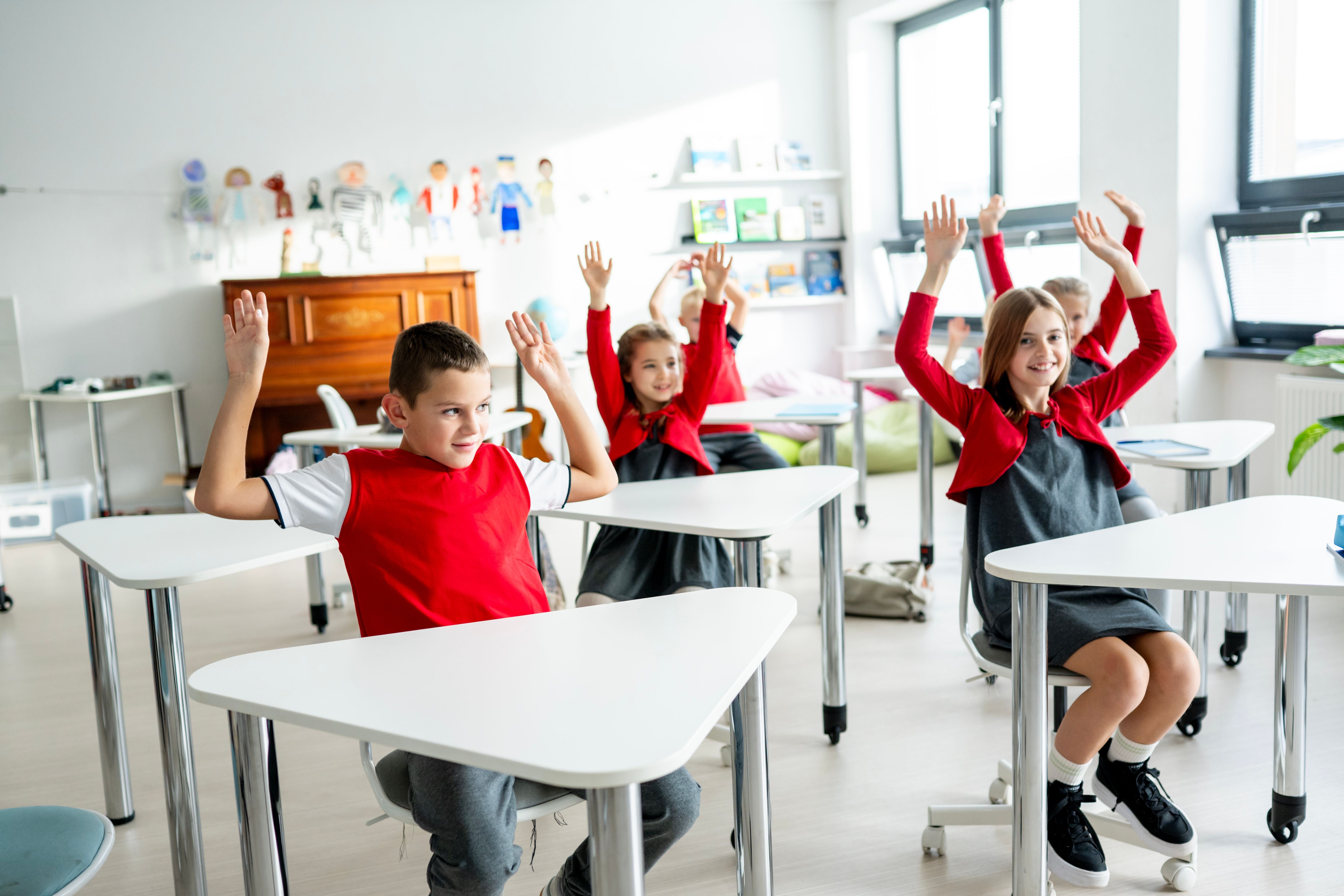 Kinder sitzen im Klassenzimmer und heben lächelnd die Hände.