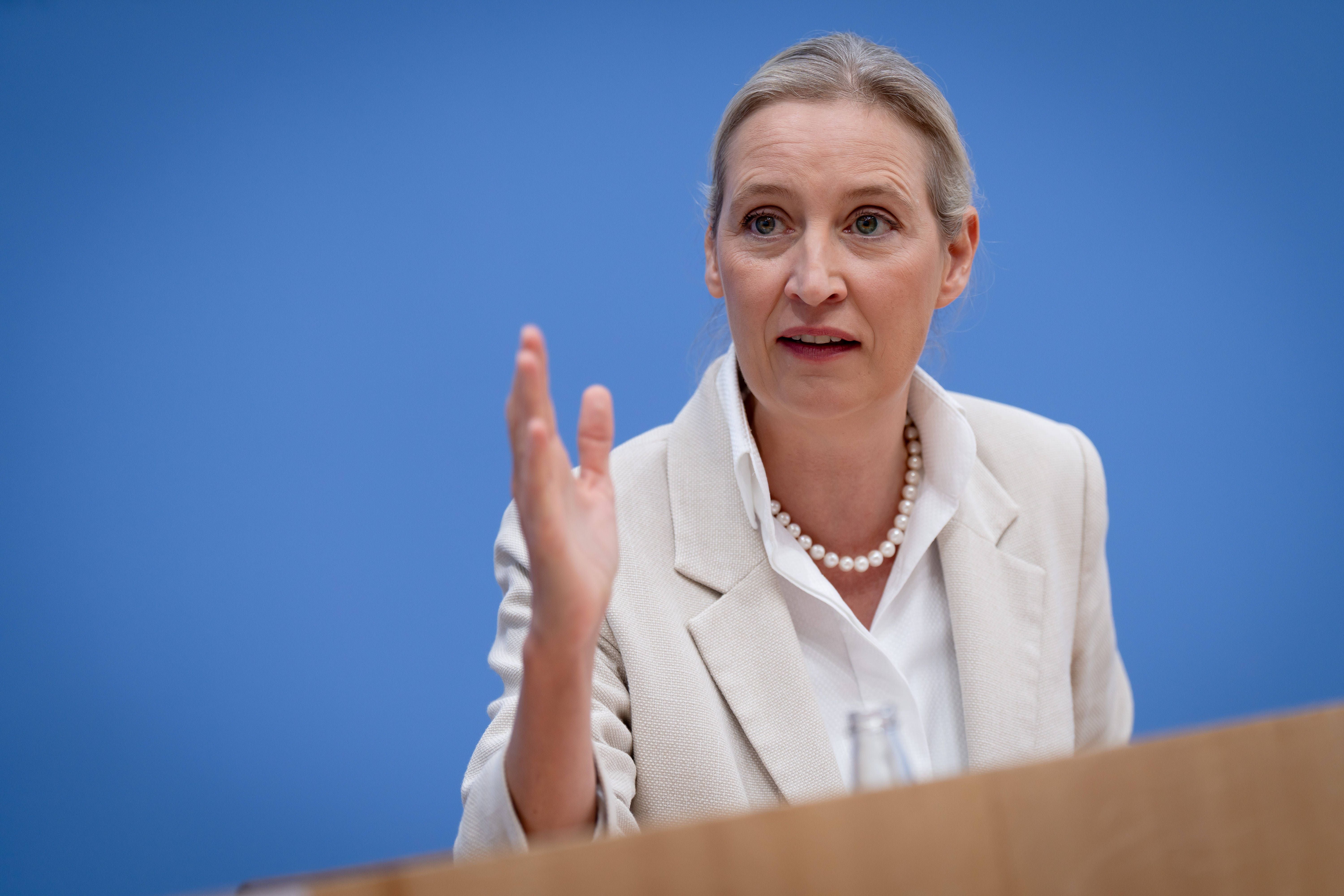 Frau im weißen Blazer spricht bei einer Pressekonferenz vor blauem Hintergrund.