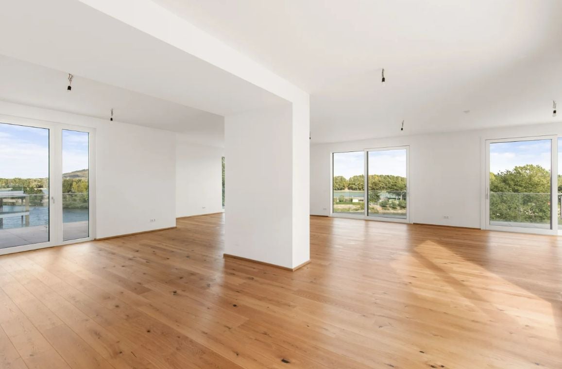 Helle, leere Wohnung mit großen Fenstern, Parkettboden und Blick ins Grüne.