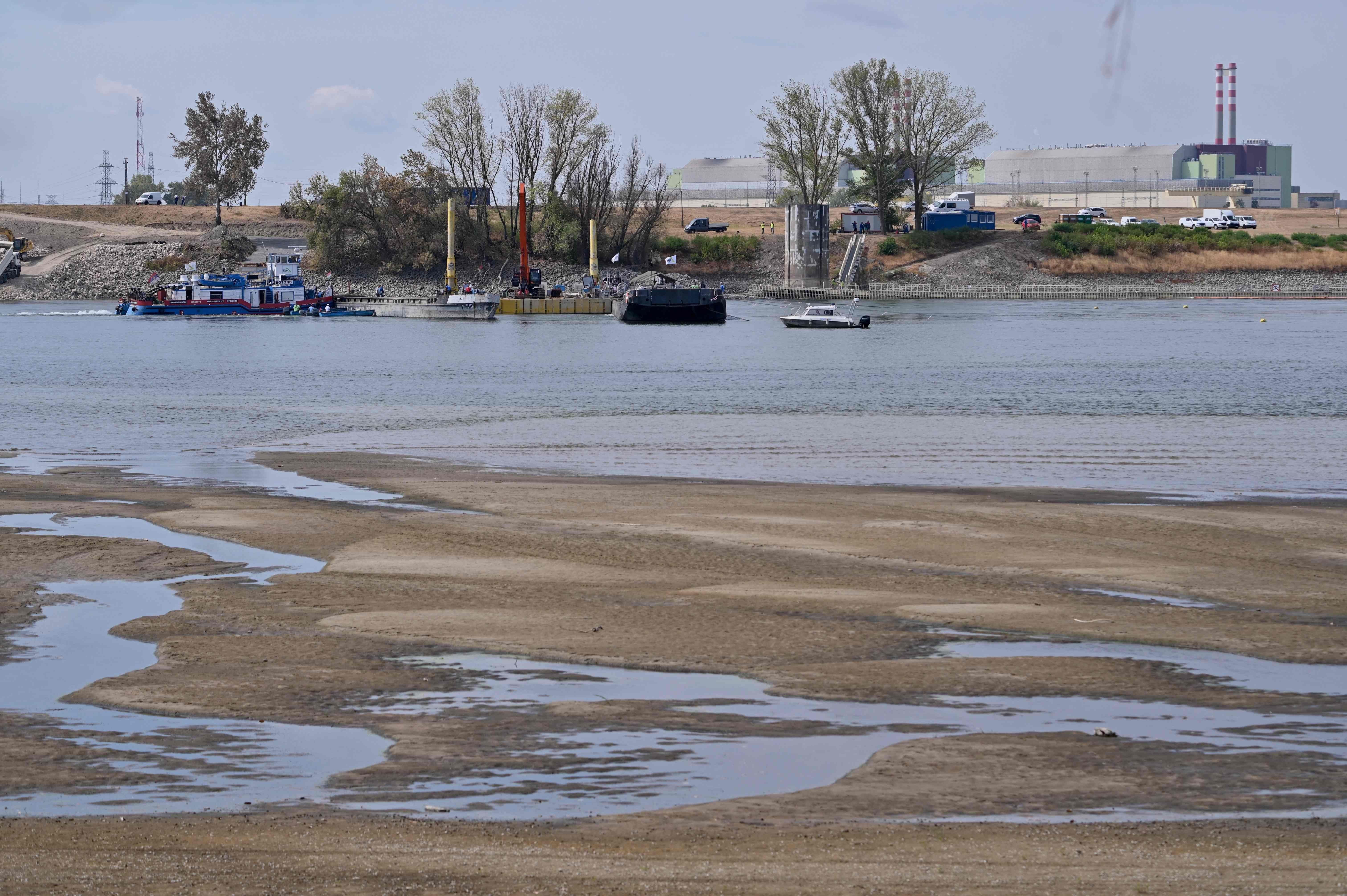 Blick auf das teils trockene Flussbett der Donau mit Schiffen und dem AKW Paks im Hintergrund.