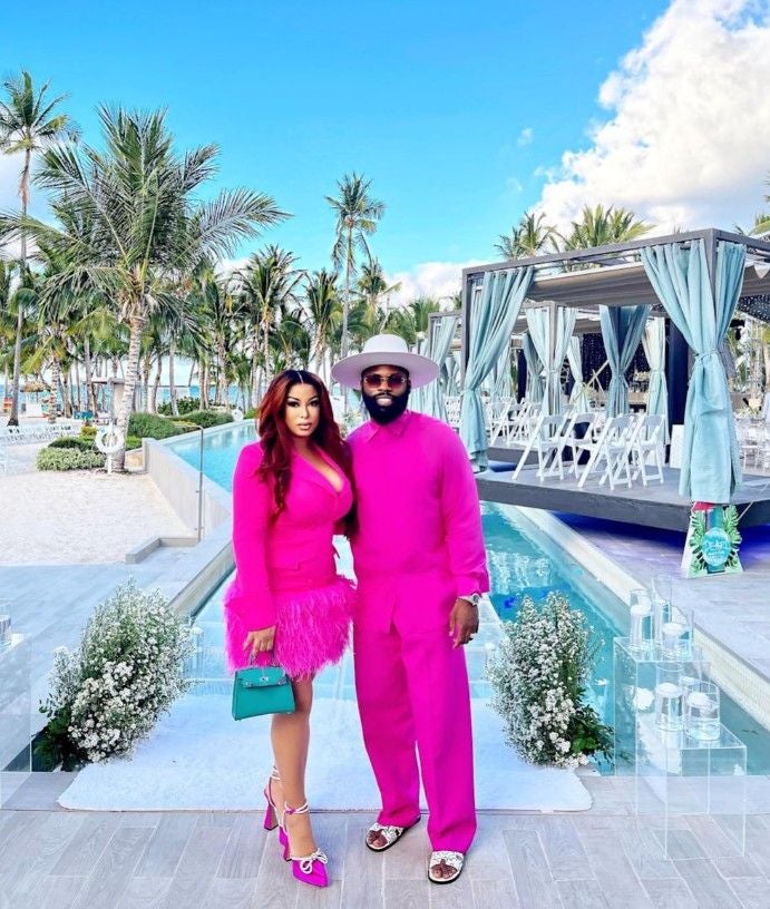Ein Paar in auffälligen pinken Outfits posiert vor einem Pool mit Palmen im Hintergrund.