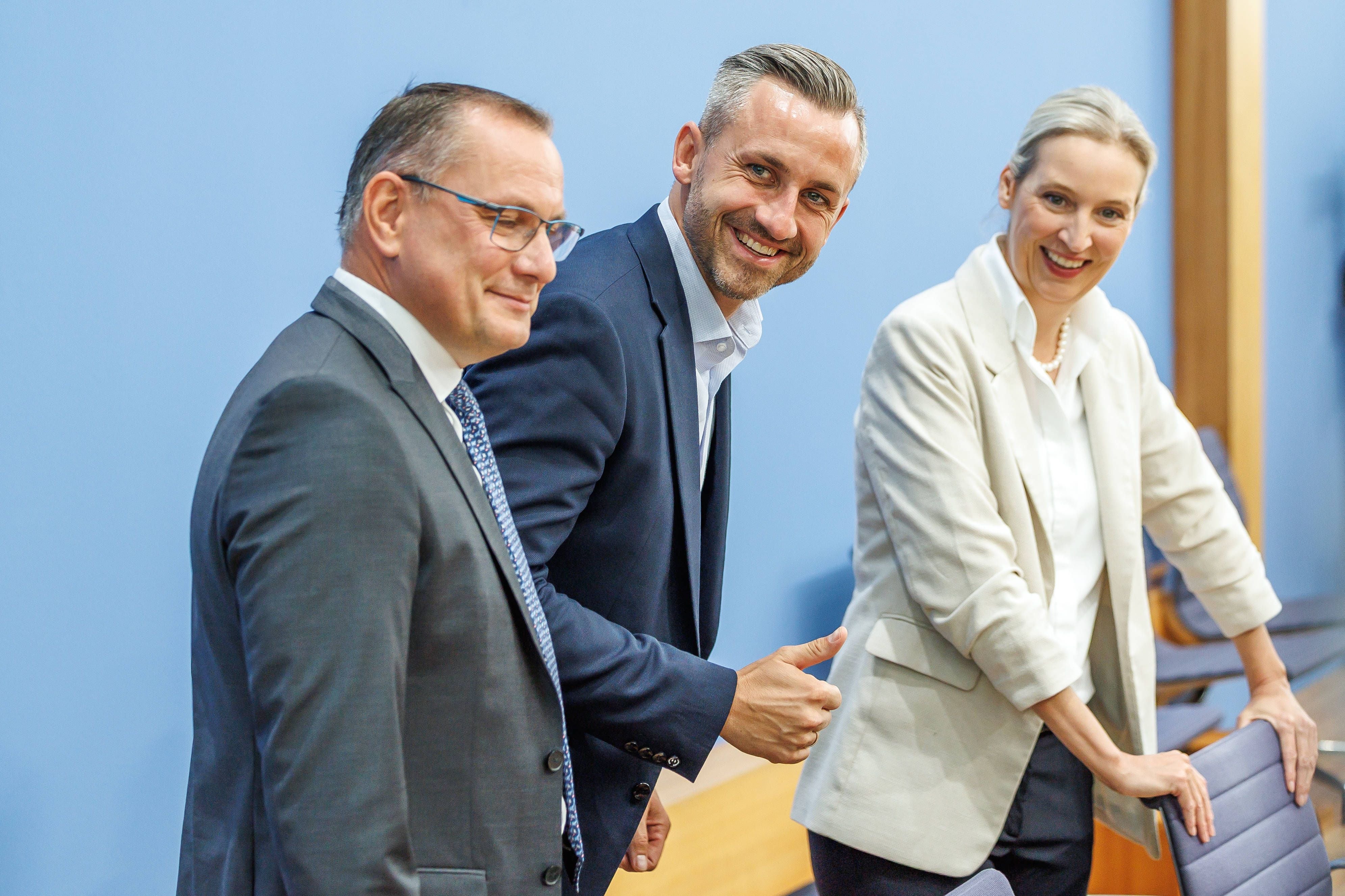Drei Personen im Anzug und Blazer stehen lächelnd nebeneinander vor einer blauen Wand.