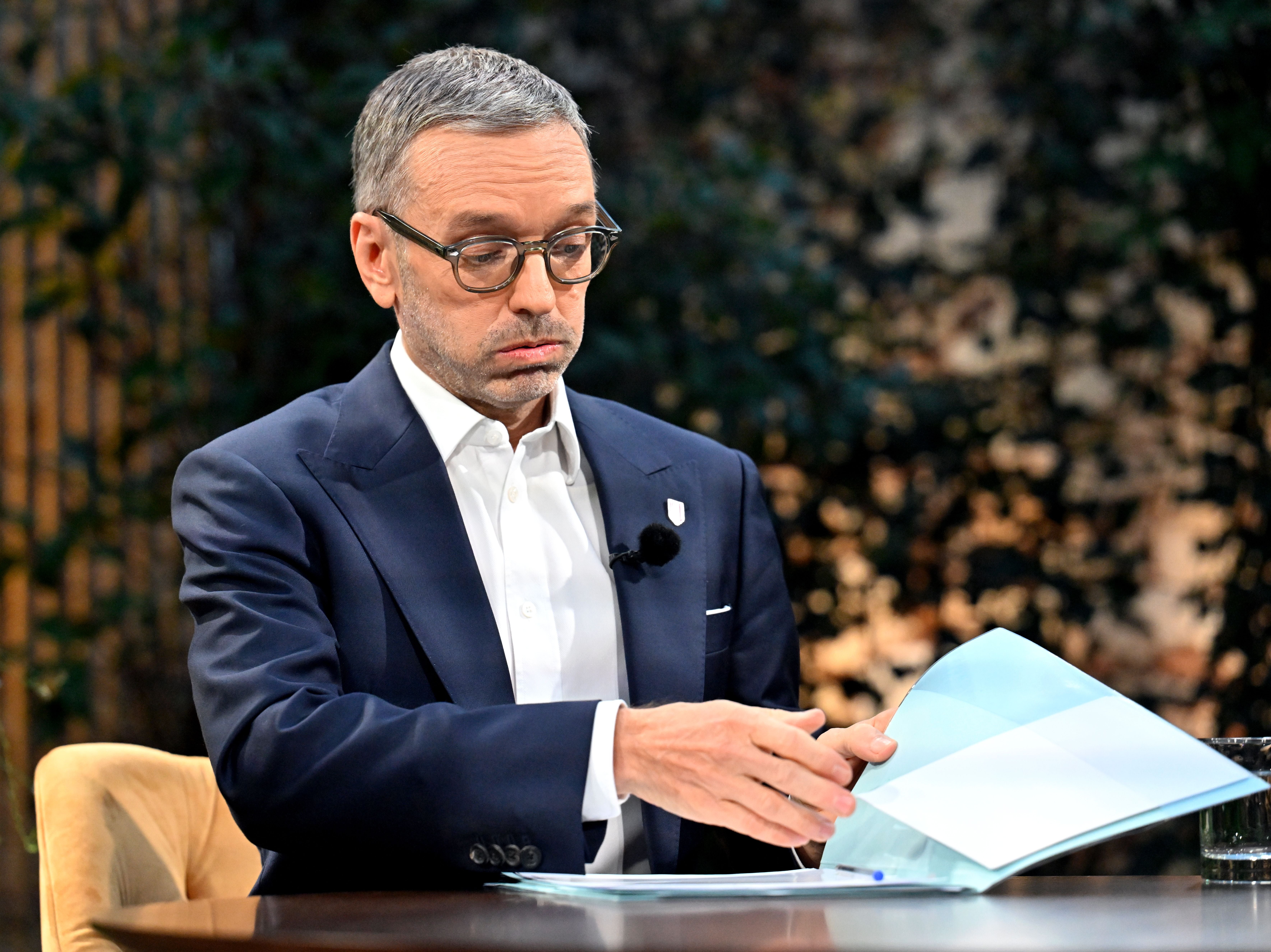 Ein Mann im Anzug blättert in Dokumenten und sitzt an einem Tisch mit Mikrofon am Revers.