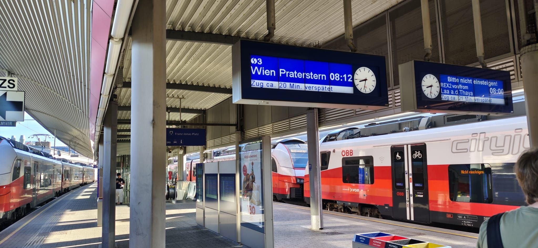 Bahnhof mit ÖBB-Zügen, Anzeigetafeln für Wien Praterstern und Verspätung, Reisende am Bahnsteig.