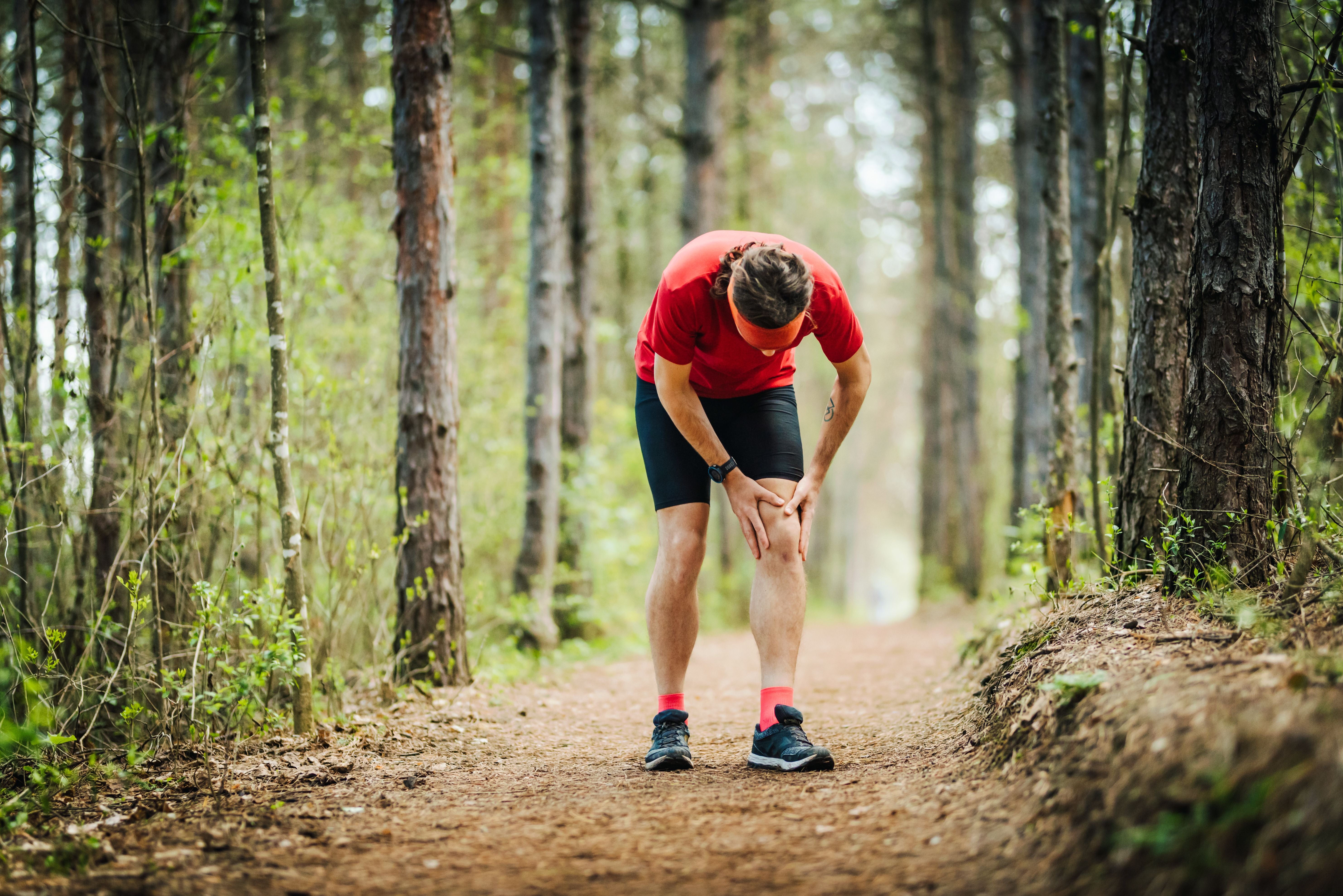 Junger Mann hält sich beim Joggen im Wald schmerzhaft das Knie.