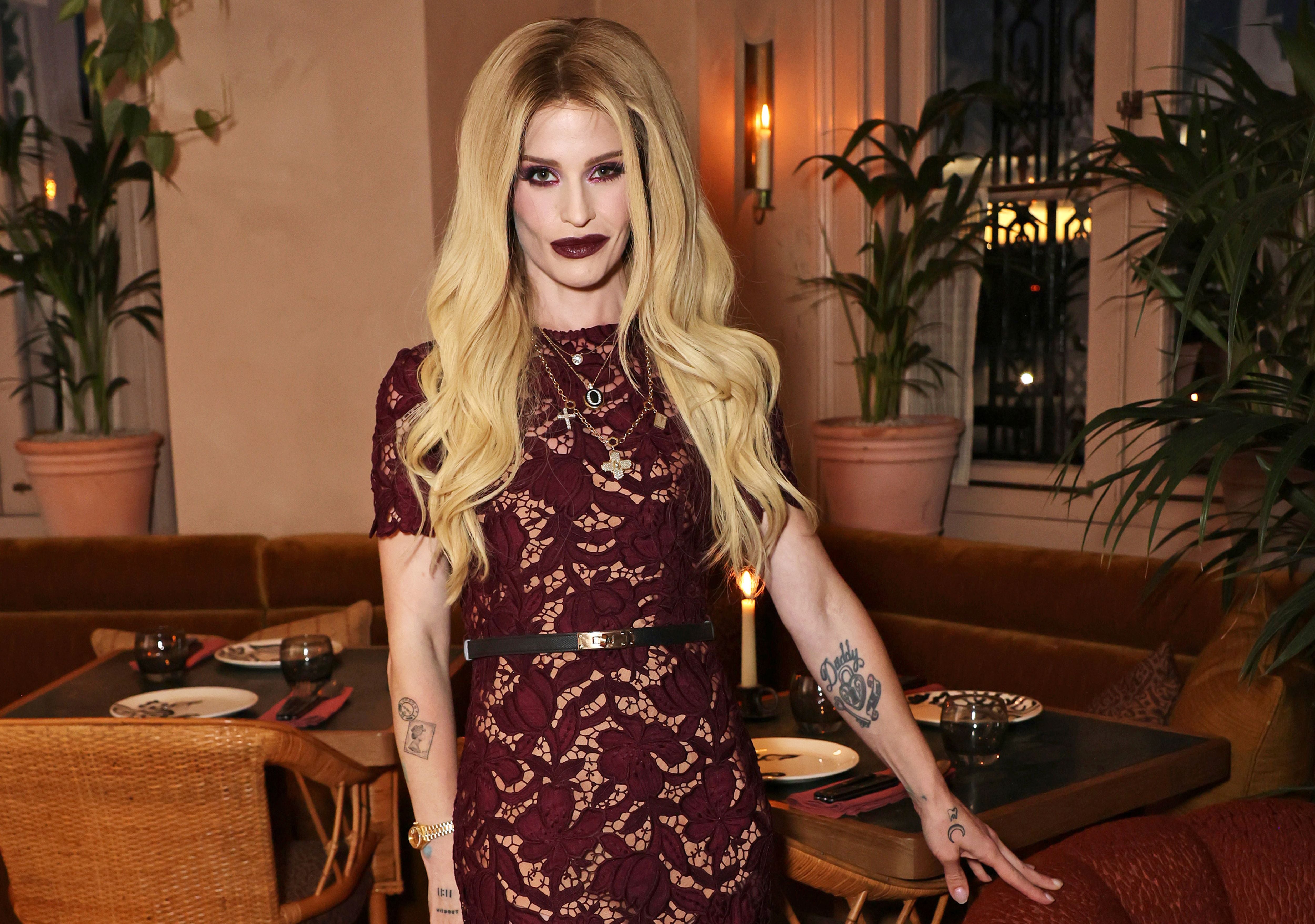Kelly Osbourne mit langen blonden Haaren und Spitzenkleid steht in stilvollem Restaurant mit Pflanzen.