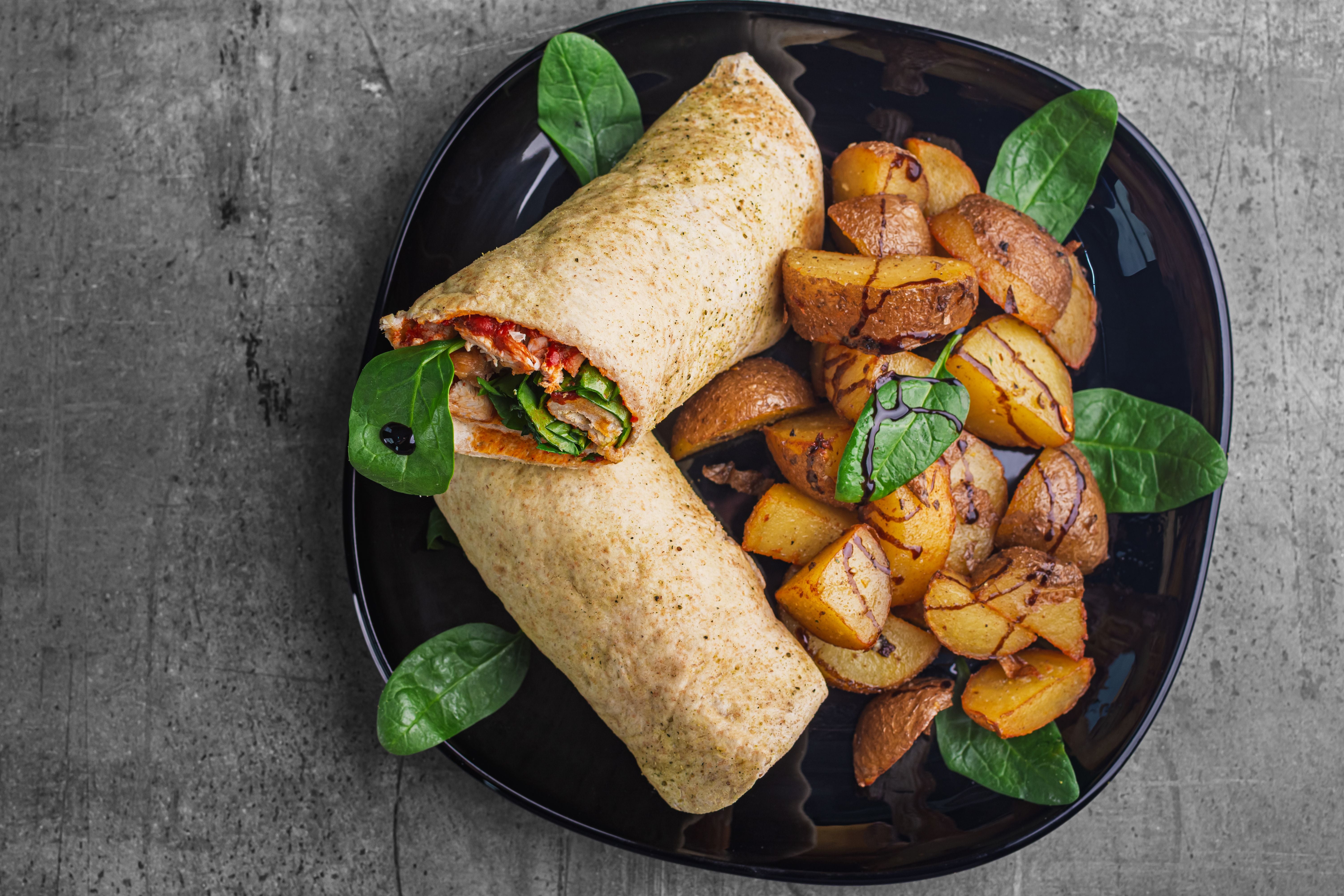 Hähnchen-Tortilla-Wrap mit Bratkartoffeln und frischem Spinat auf schwarzem Teller.
