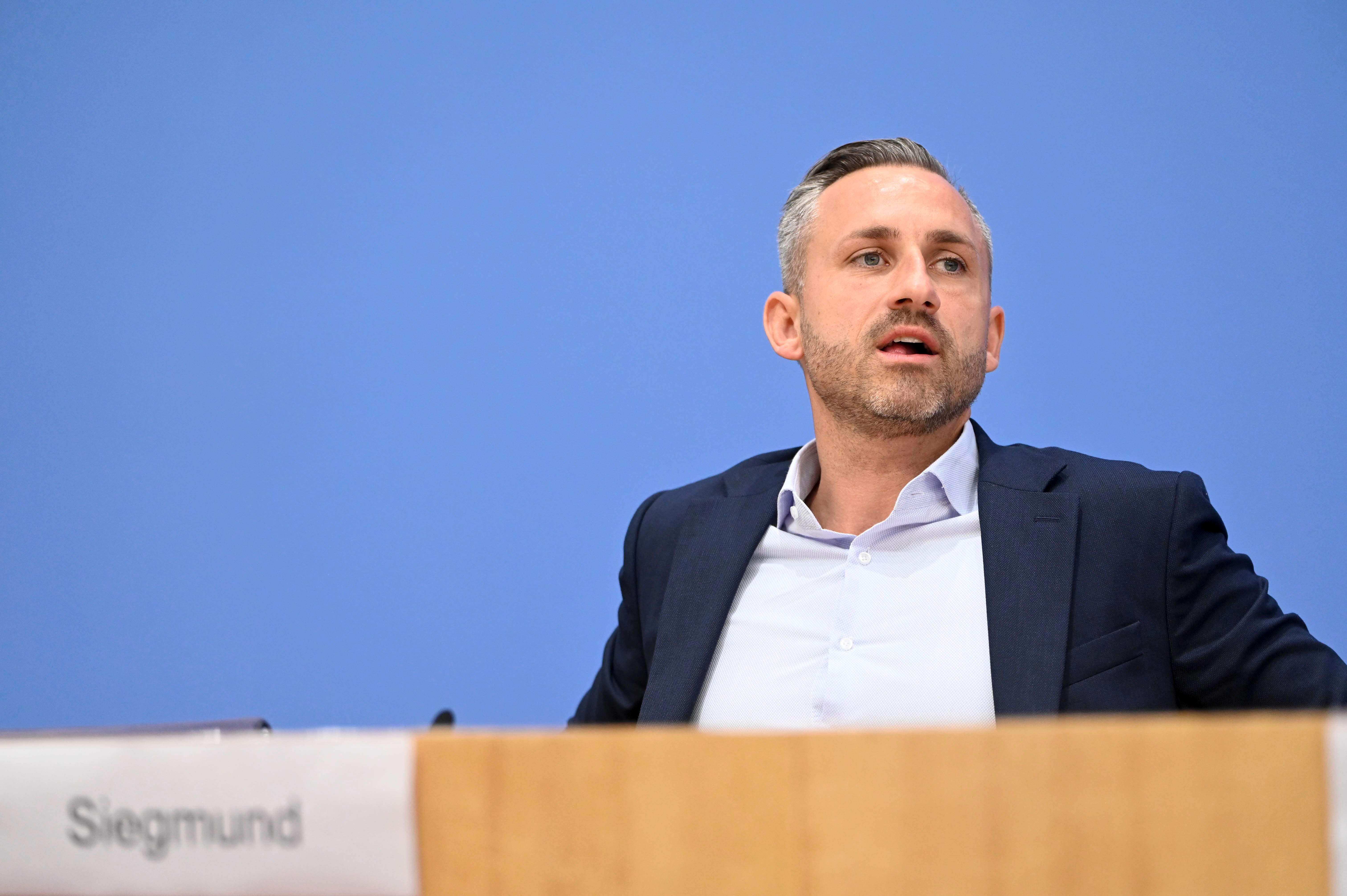 Ein Mann im Anzug spricht bei einer Pressekonferenz vor blauem Hintergrund.