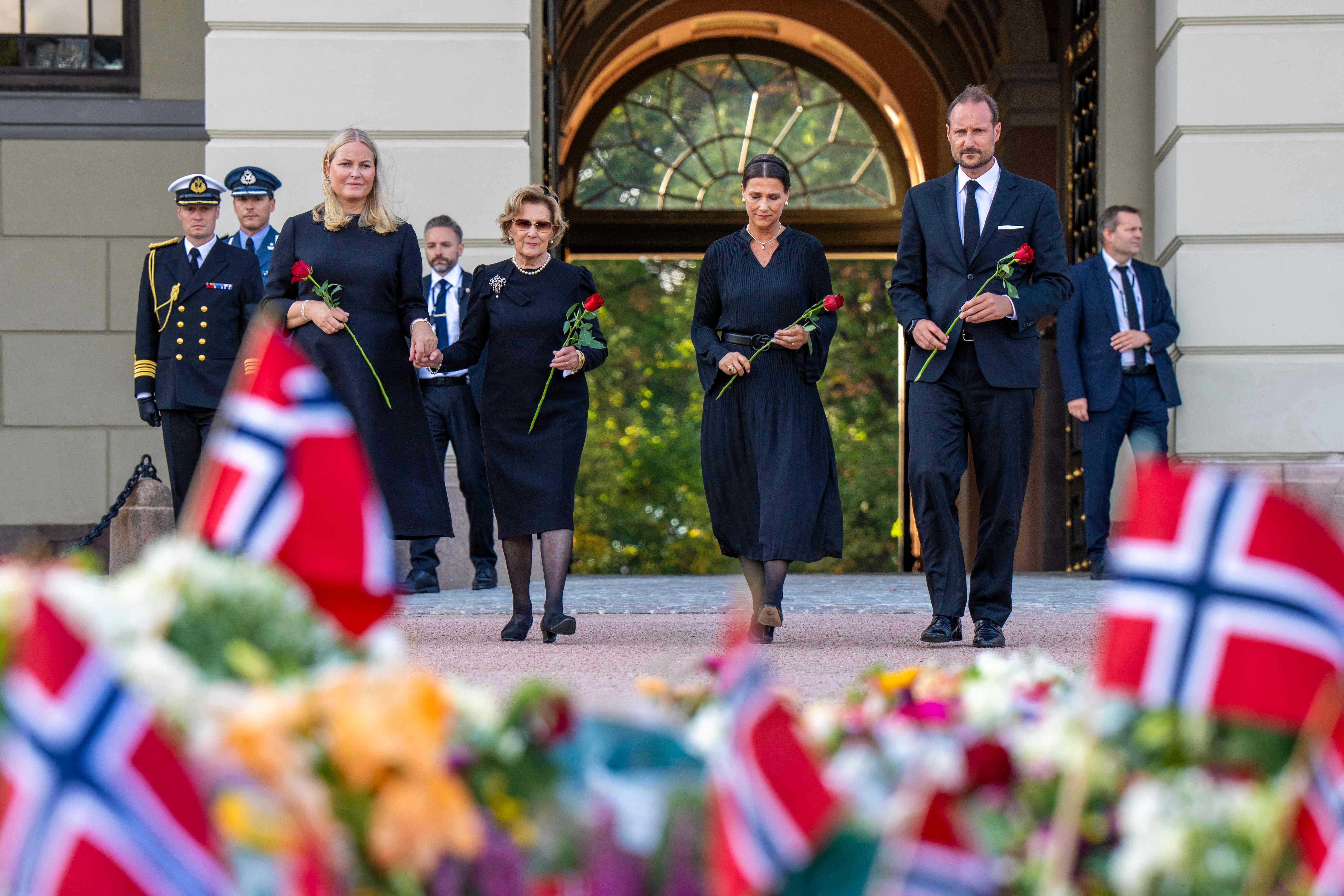 Vier Personen in schwarzer Kleidung halten Rosen, im Vordergrund Blumen und norwegische Flaggen.