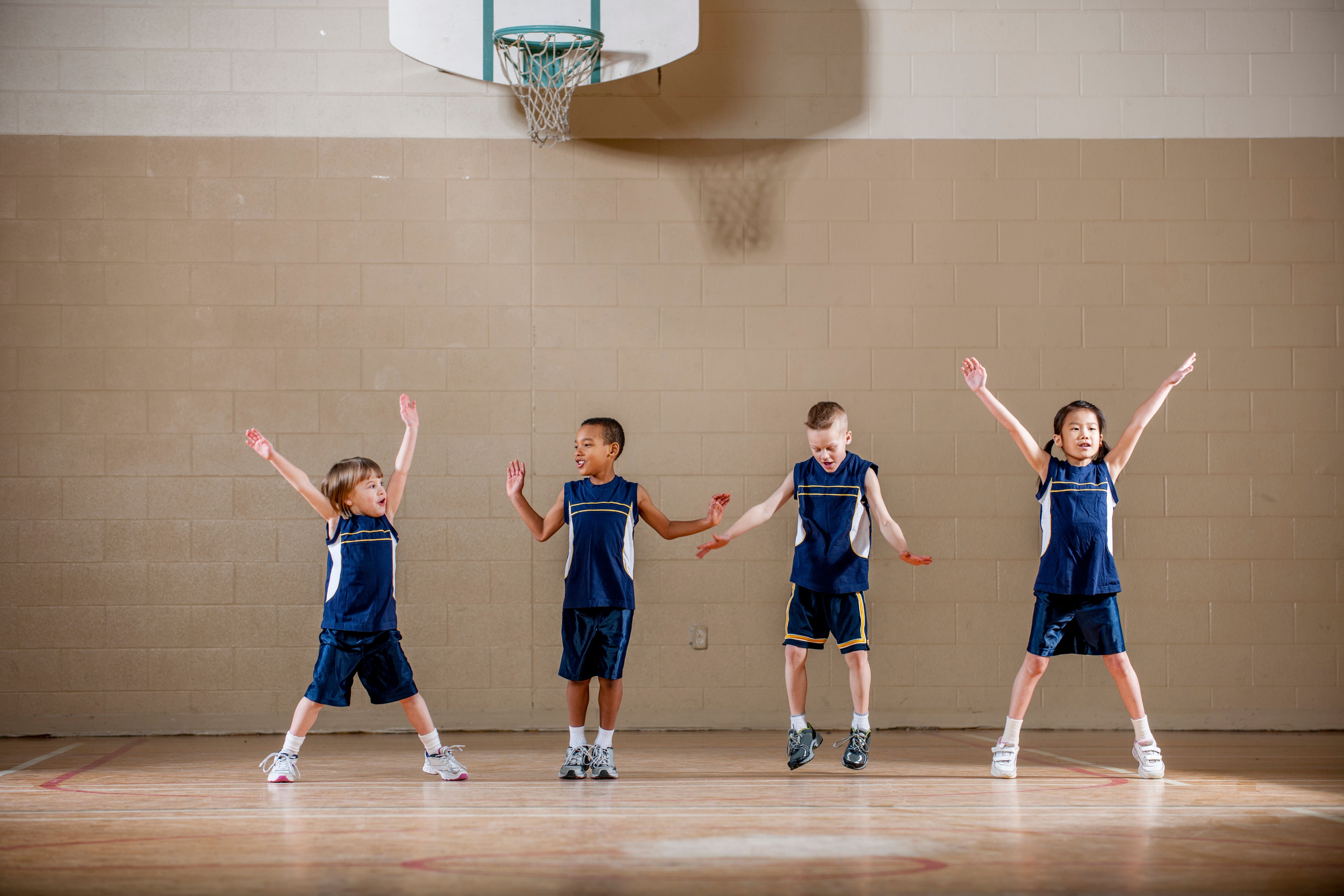 Vier Kinder in blauen Trikots machen Hampelmänner in einer Turnhalle unter einem Basketballkorb.