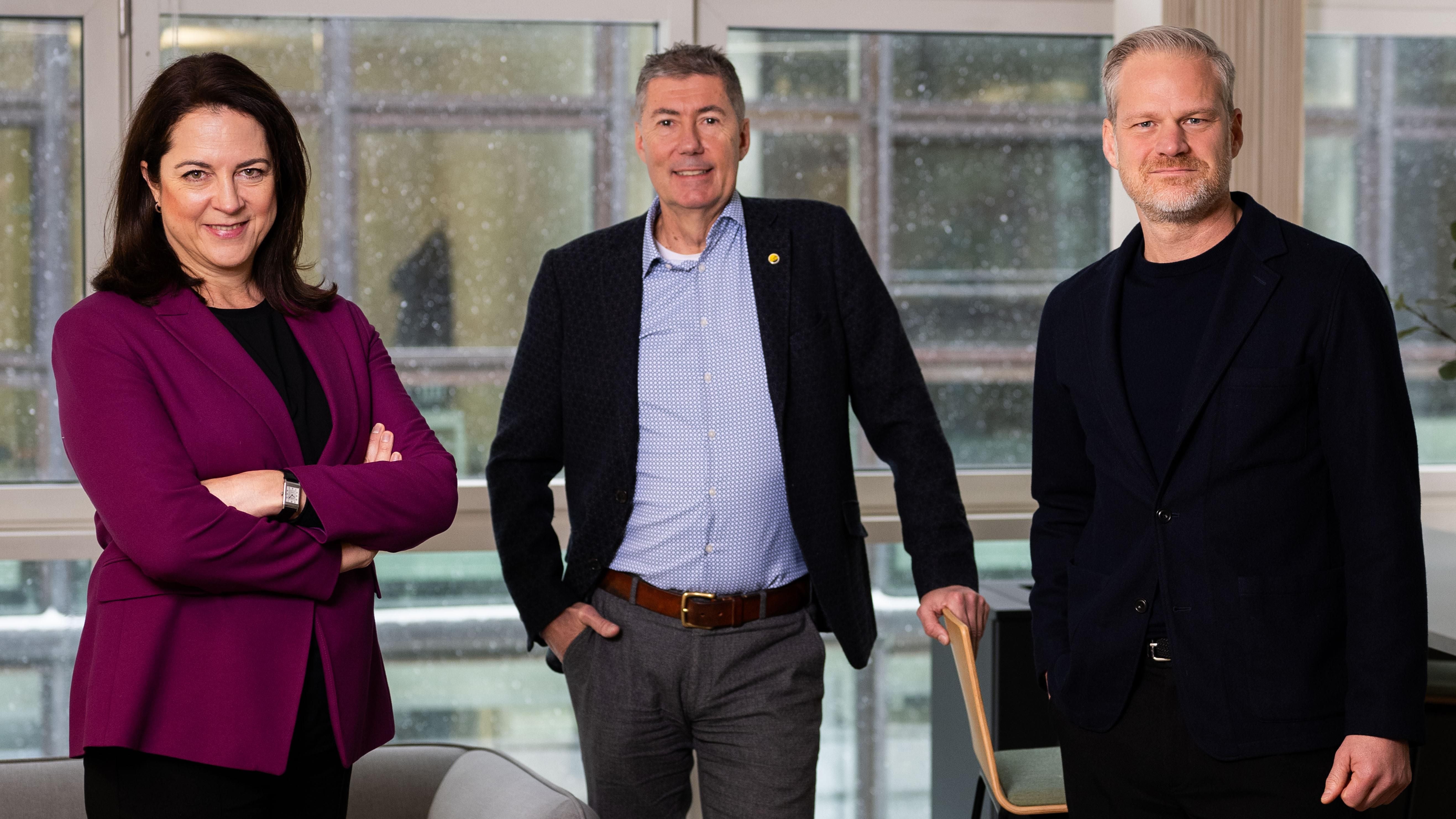 Drei Personen posieren gemeinsam vor Fenstern, alle in Businesskleidung.