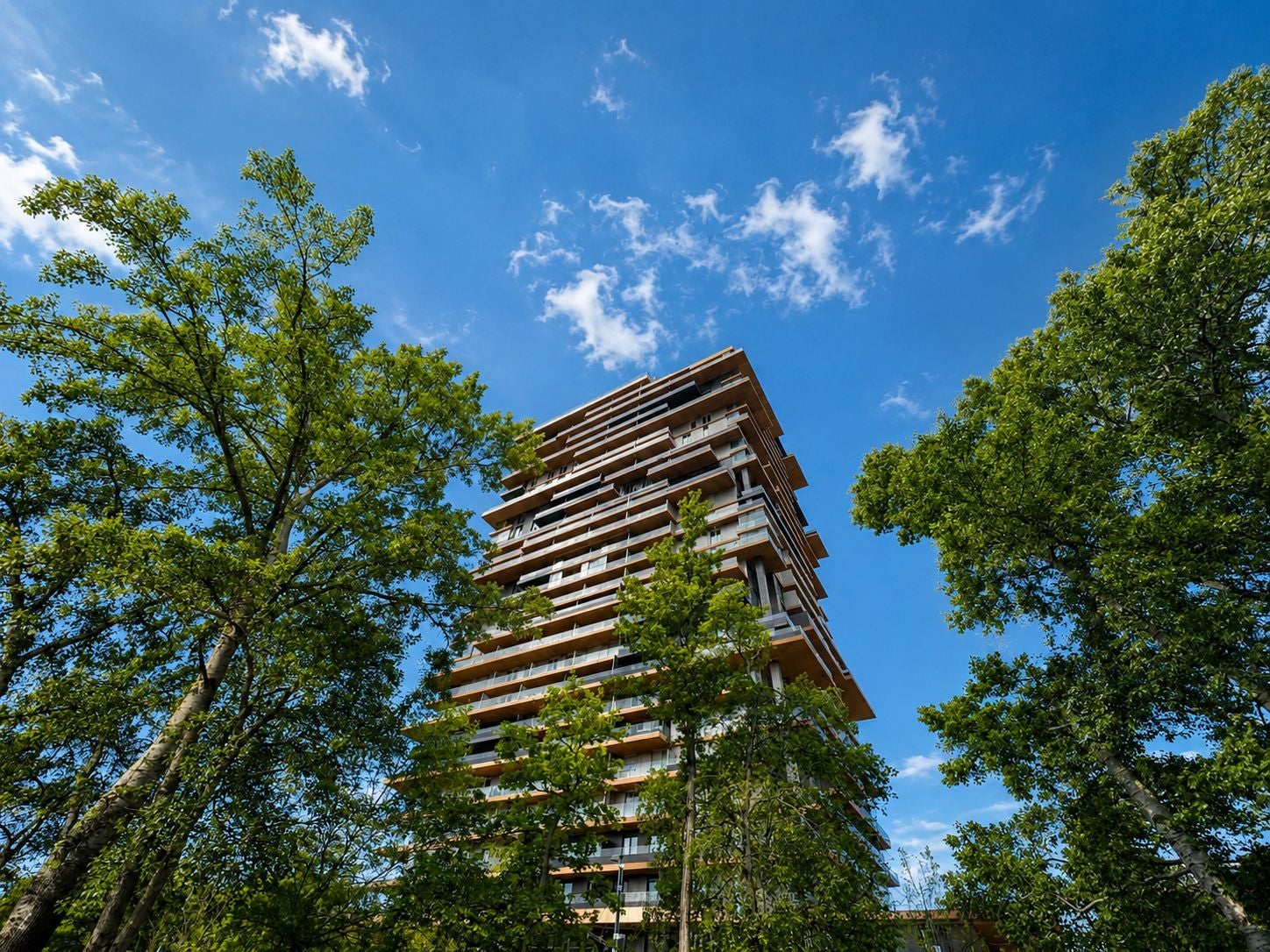 Modernes Hochhaus zwischen grünen Bäumen vor blauem Himmel mit wenigen Wolken.
