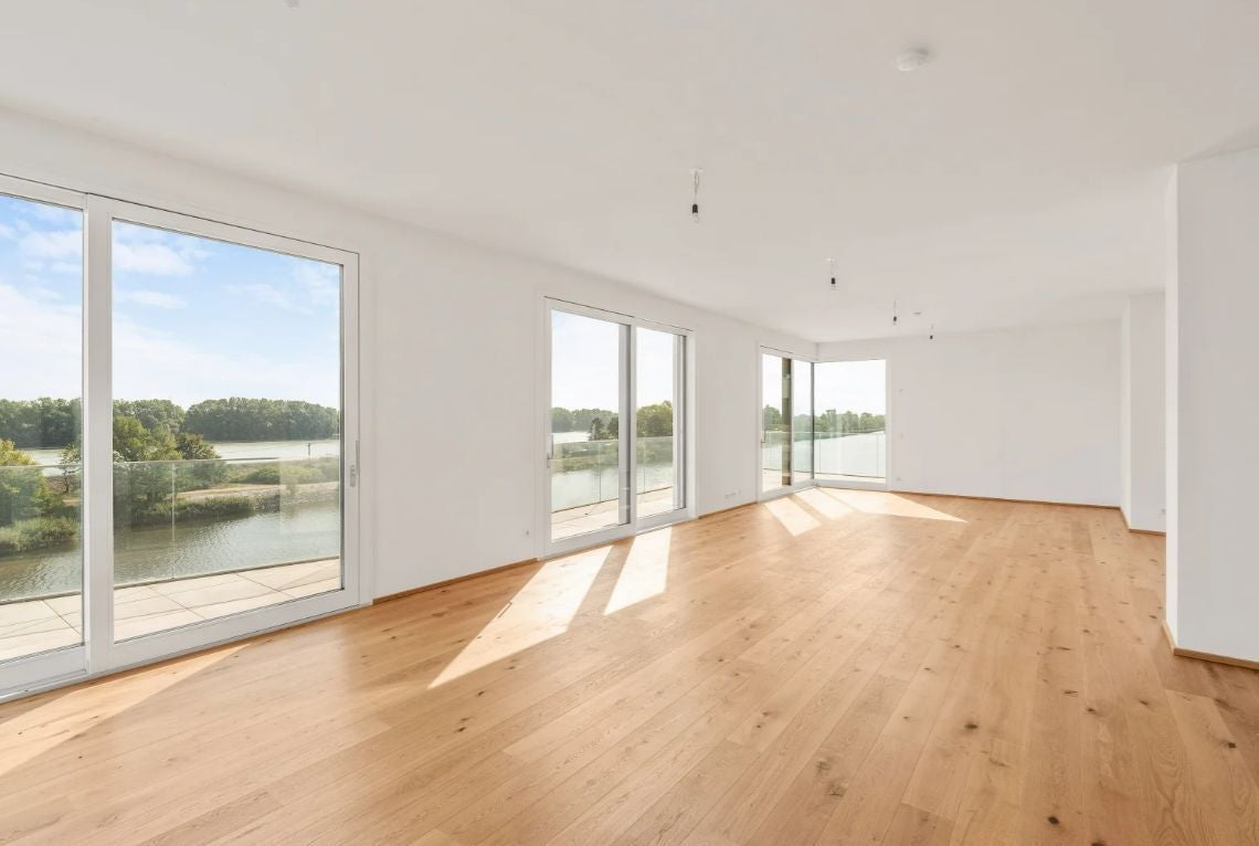 Heller, leerer Raum mit Holzfußboden und großen Fenstern mit Blick auf einen Fluss.