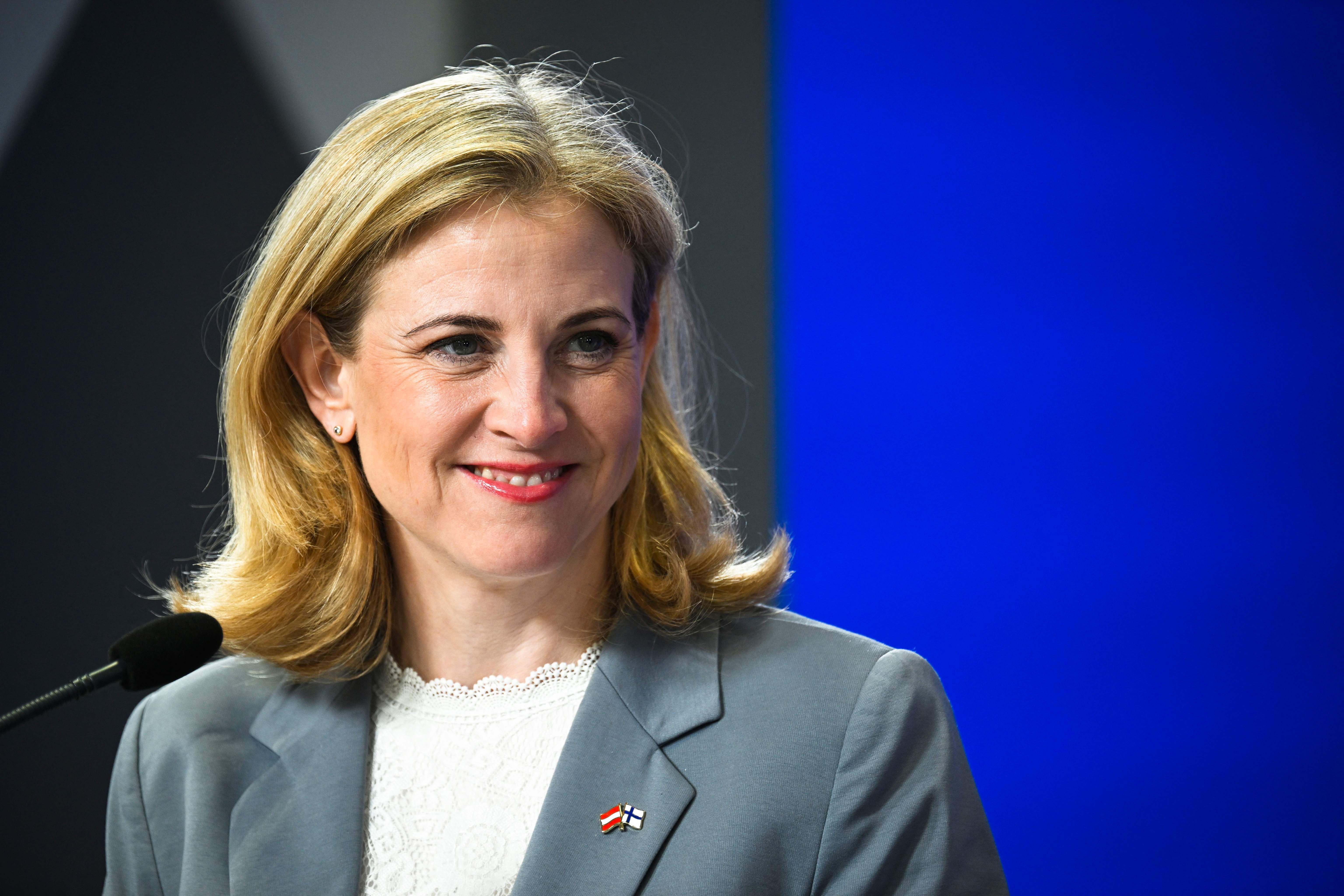 Eine Frau im grauen Blazer spricht lächelnd bei einer Pressekonferenz vor blauem Hintergrund.
