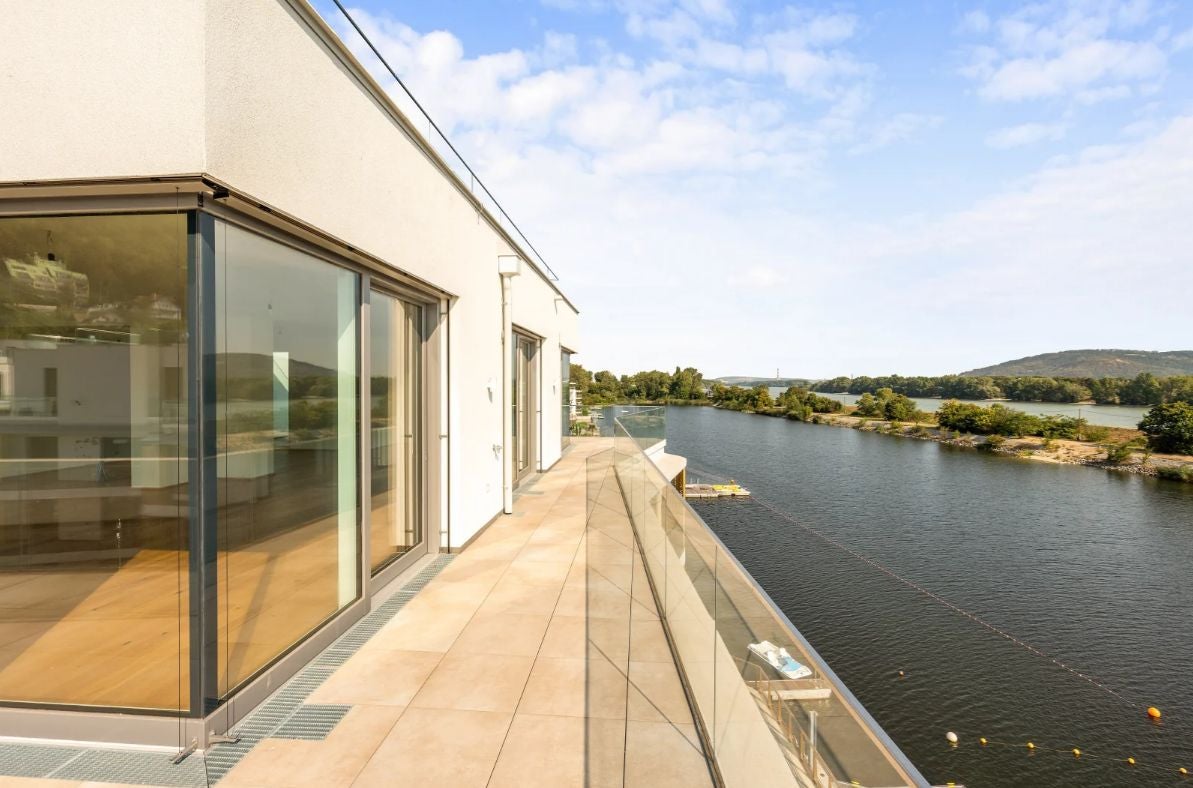 Moderne Terrasse mit Glasgeländer und Blick auf einen Fluss mit bewaldetem Ufer.