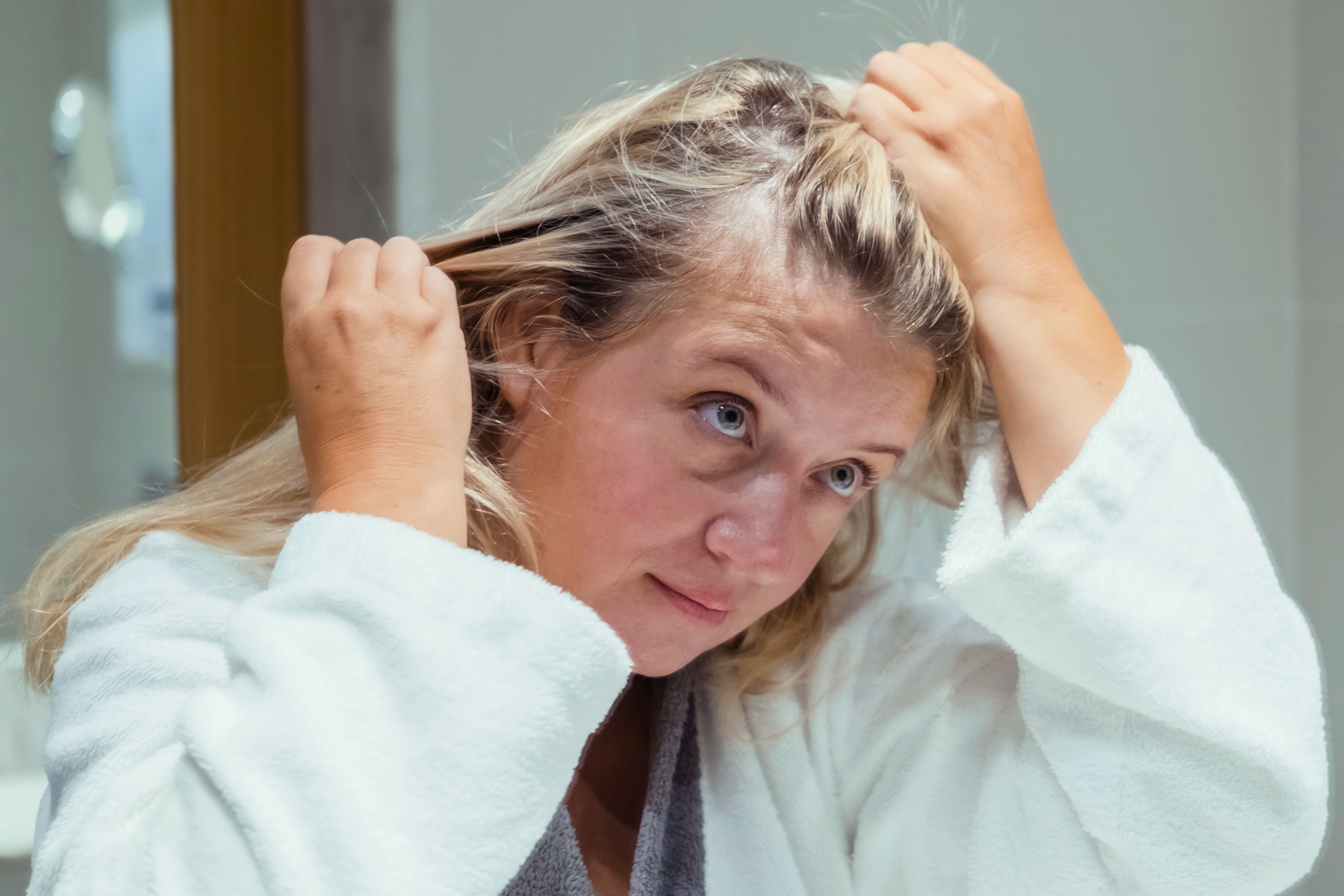 Blonde Frau massiert Haaröl in ihre Kopfhaut, trägt weißen Bademantel.