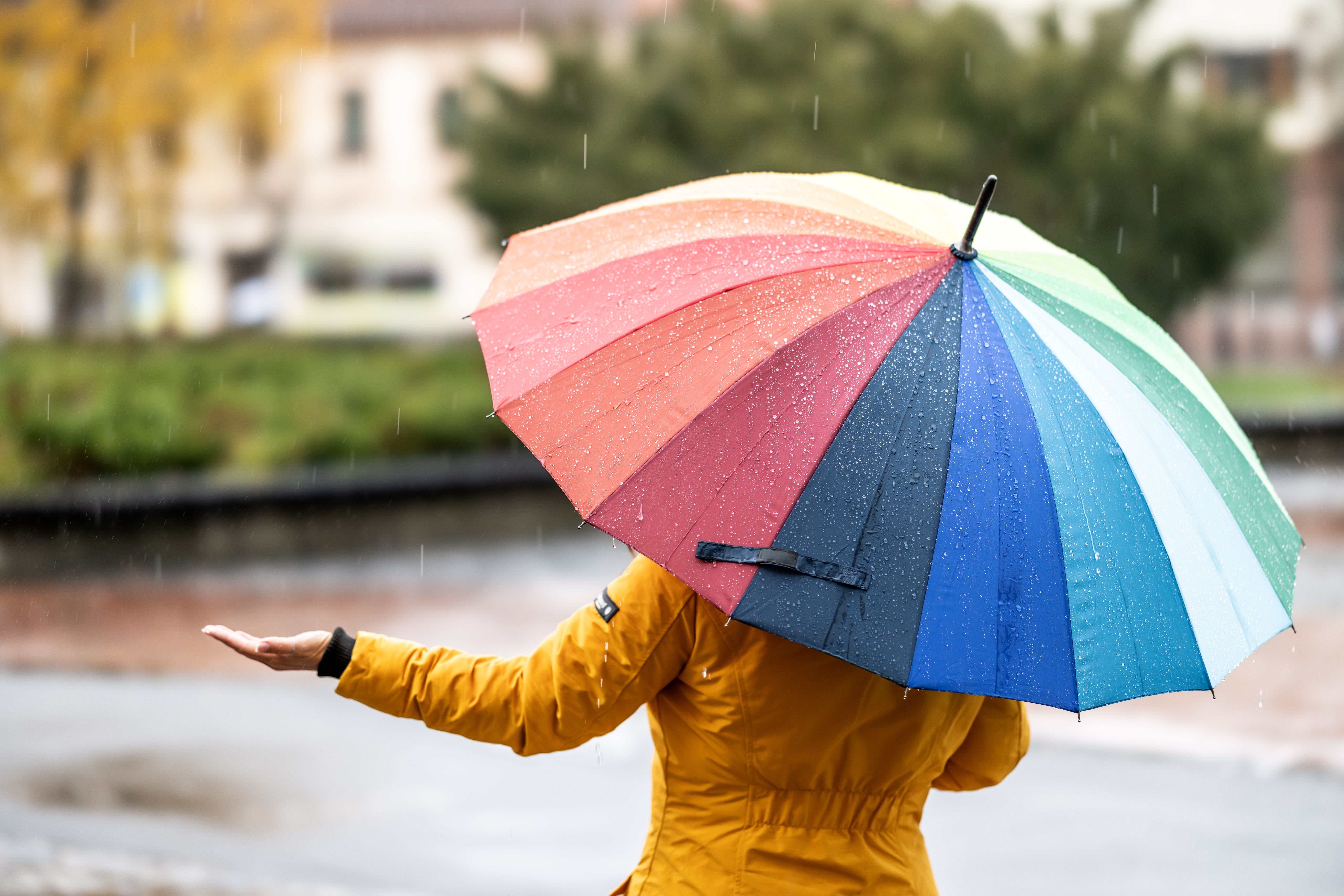Frau mit gelber Jacke hält einen bunten Regenschirm und prüft den Regen mit ausgestreckter Hand.