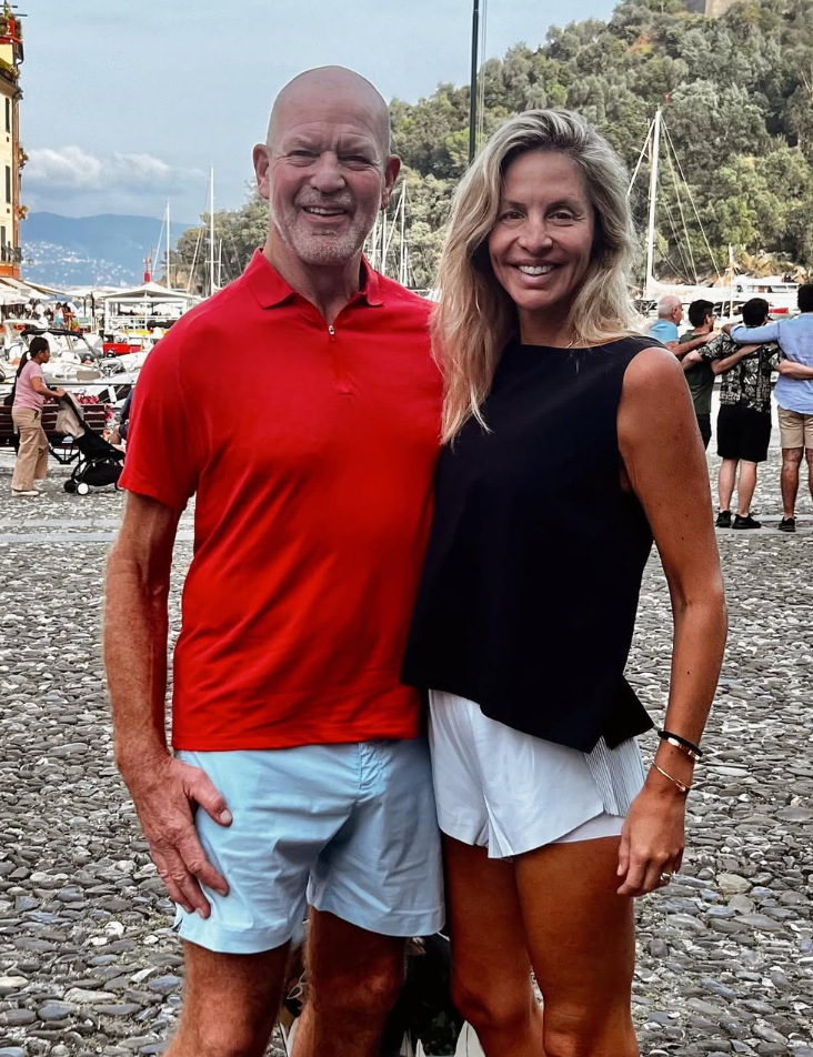 Ein Mann und eine Frau lächeln draußen auf einem gepflasterten Platz mit Yachten im Hintergrund.
