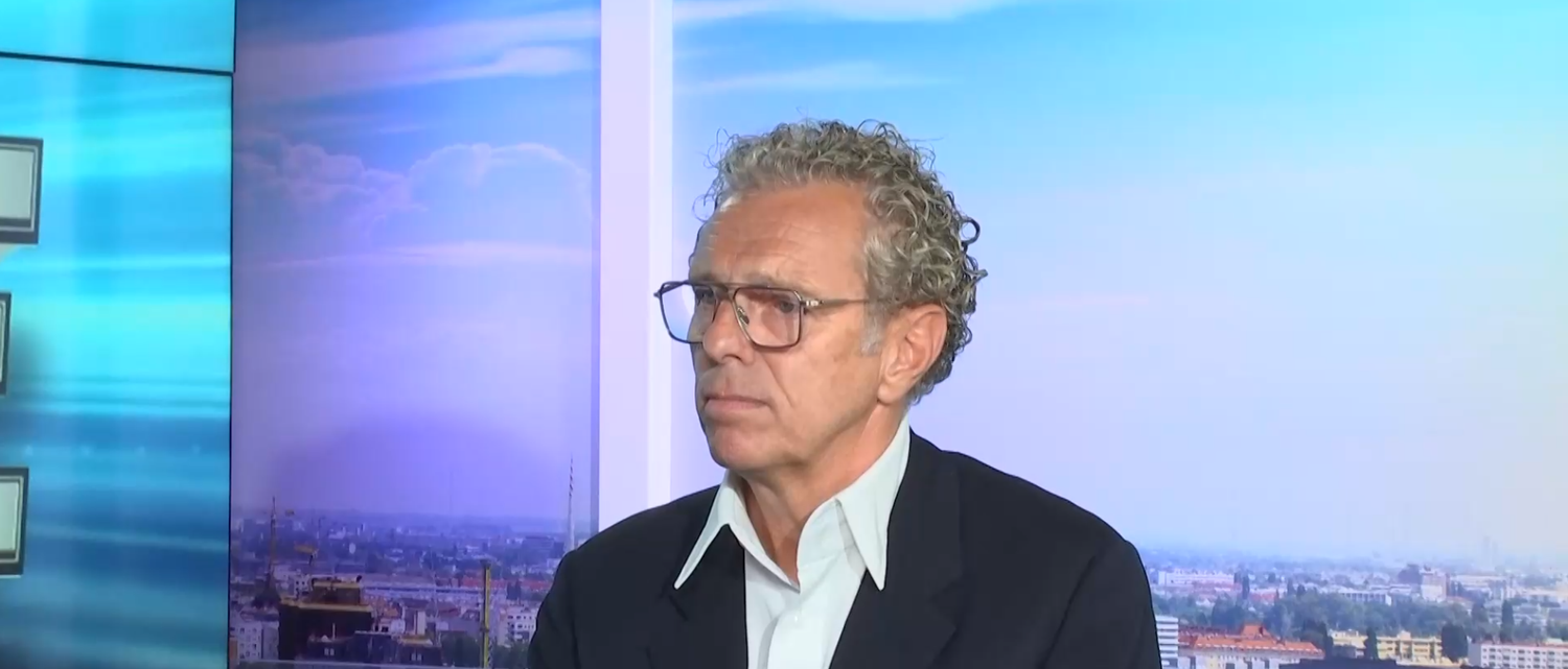 Ein Mann mit Brille und lockigem Haar sitzt vor einem Stadtpanorama im Studio.