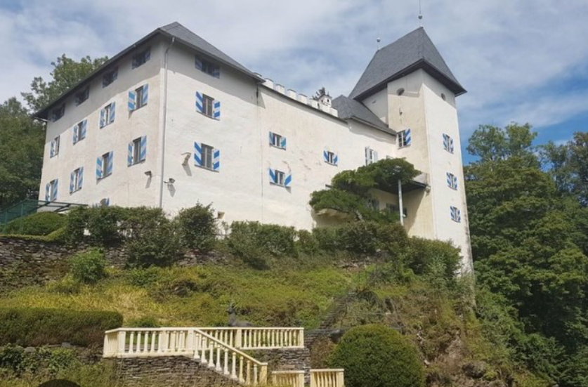 Weißes Schloss Drasing mit blauen Fensterläden auf einem bewaldeten Hügel unter blauem Himmel.