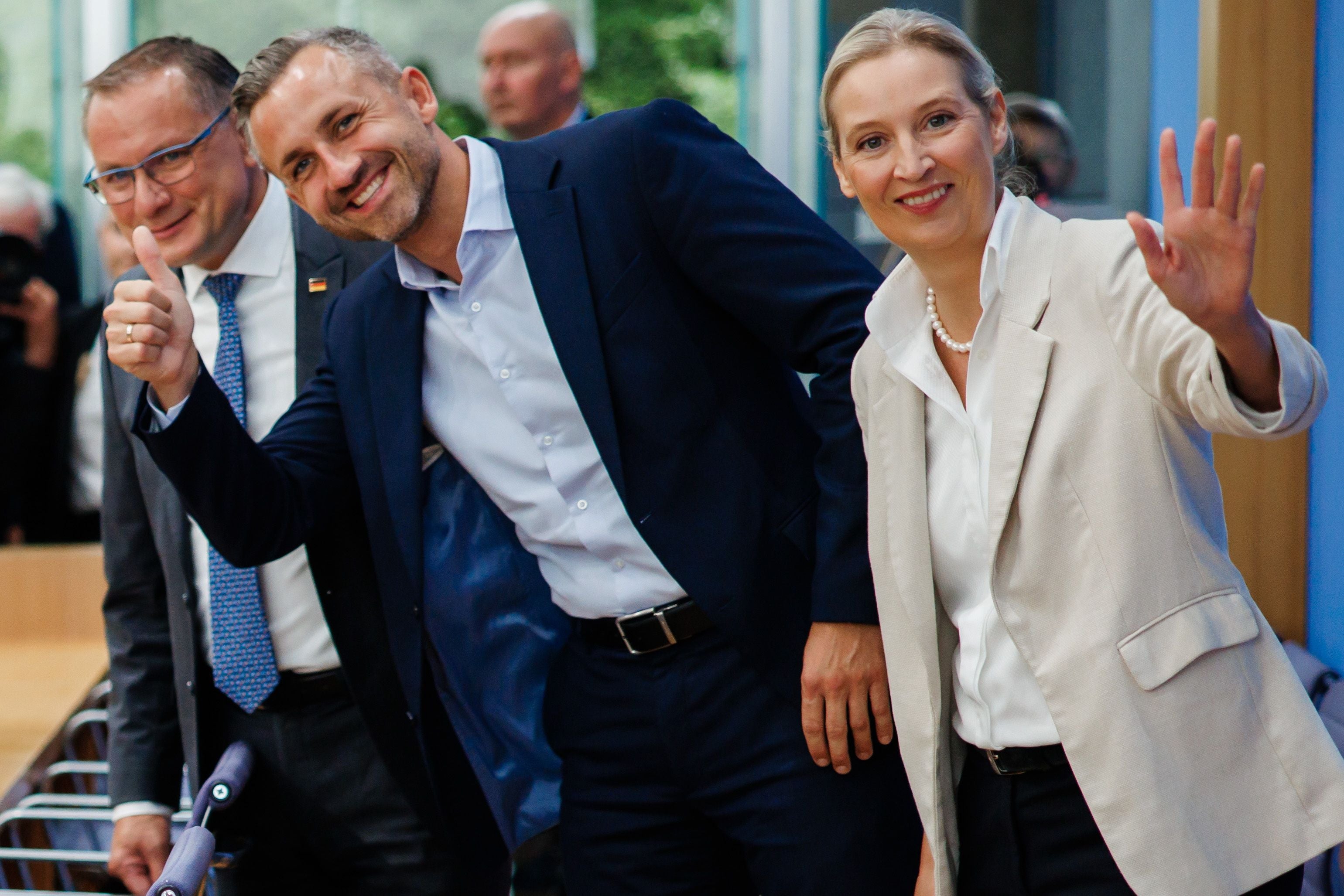 Drei Personen in Anzügen lächeln und winken bei einer Pressekonferenz.