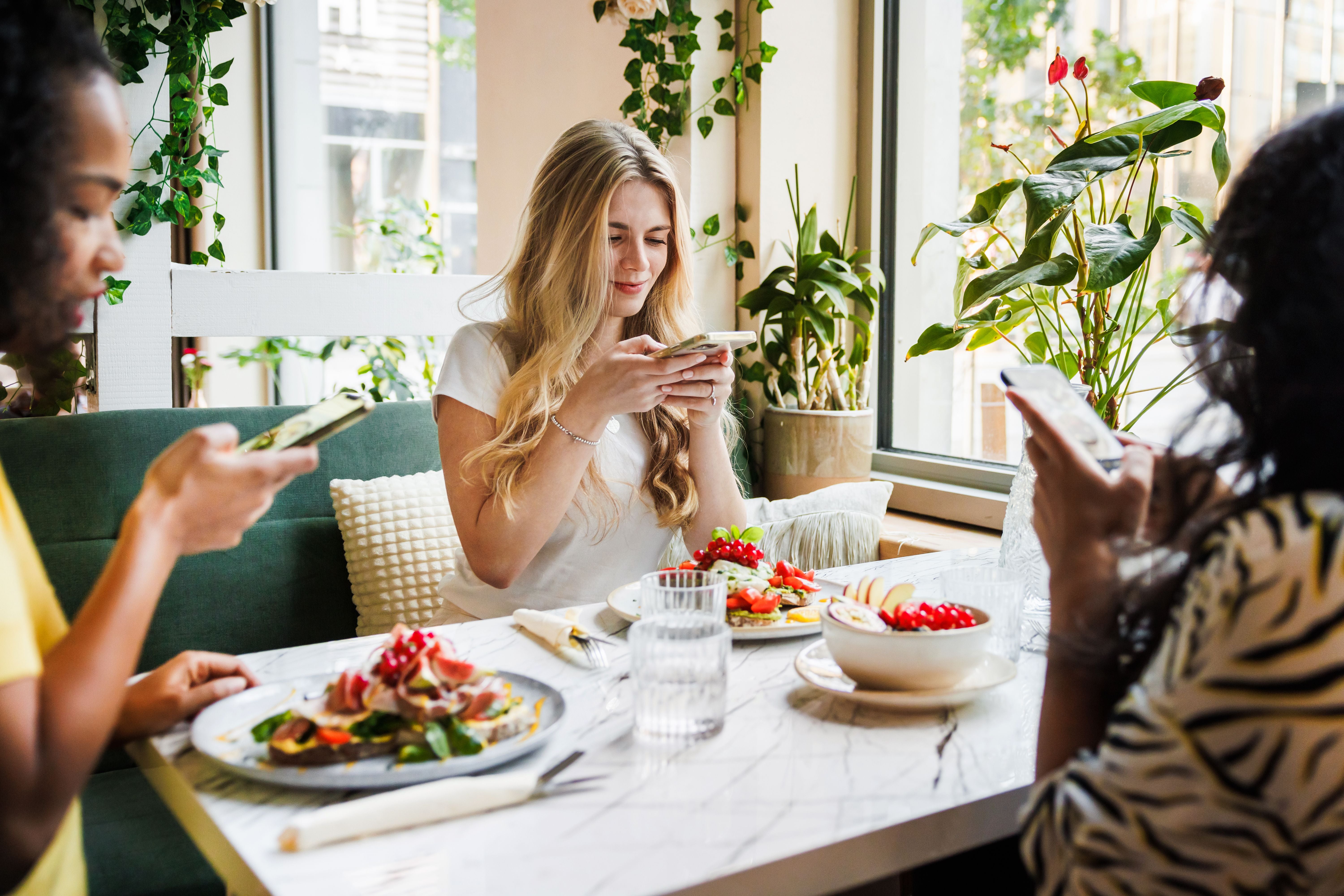 Drei Freunde sitzen im Café, essen Salat und Obst, und benutzen ihre Smartphones am Tisch.