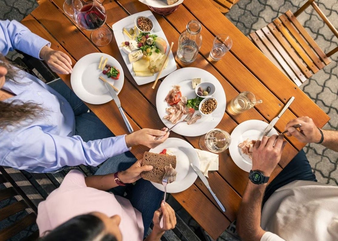 Drei Personen essen eine kalte Platte mit Brot, Wurst, Käse und Wein an einem Holztisch im Freien.