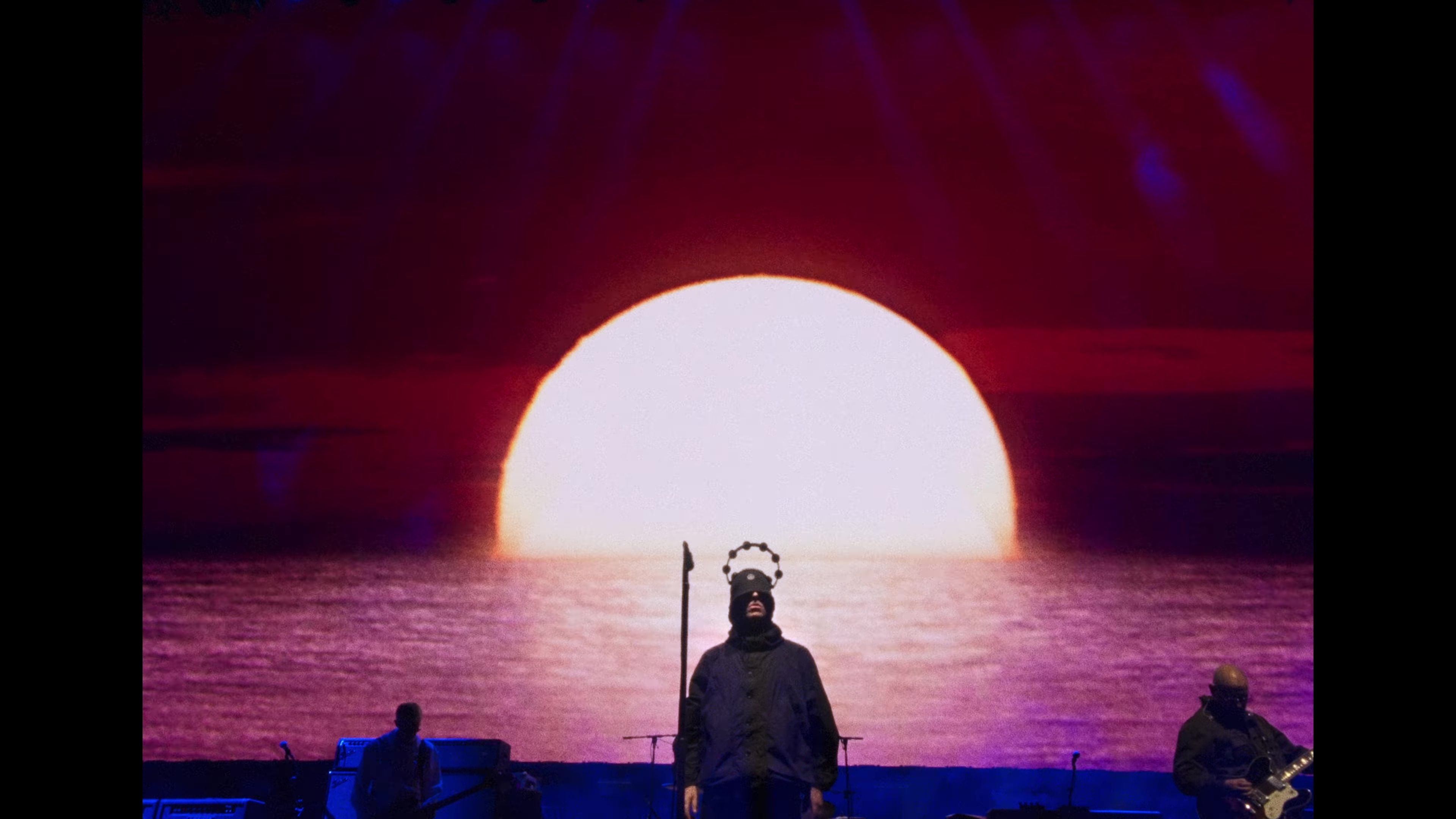 Bandmitglieder vor Bühnenbild mit großem Sonnenuntergang über dem Meer.