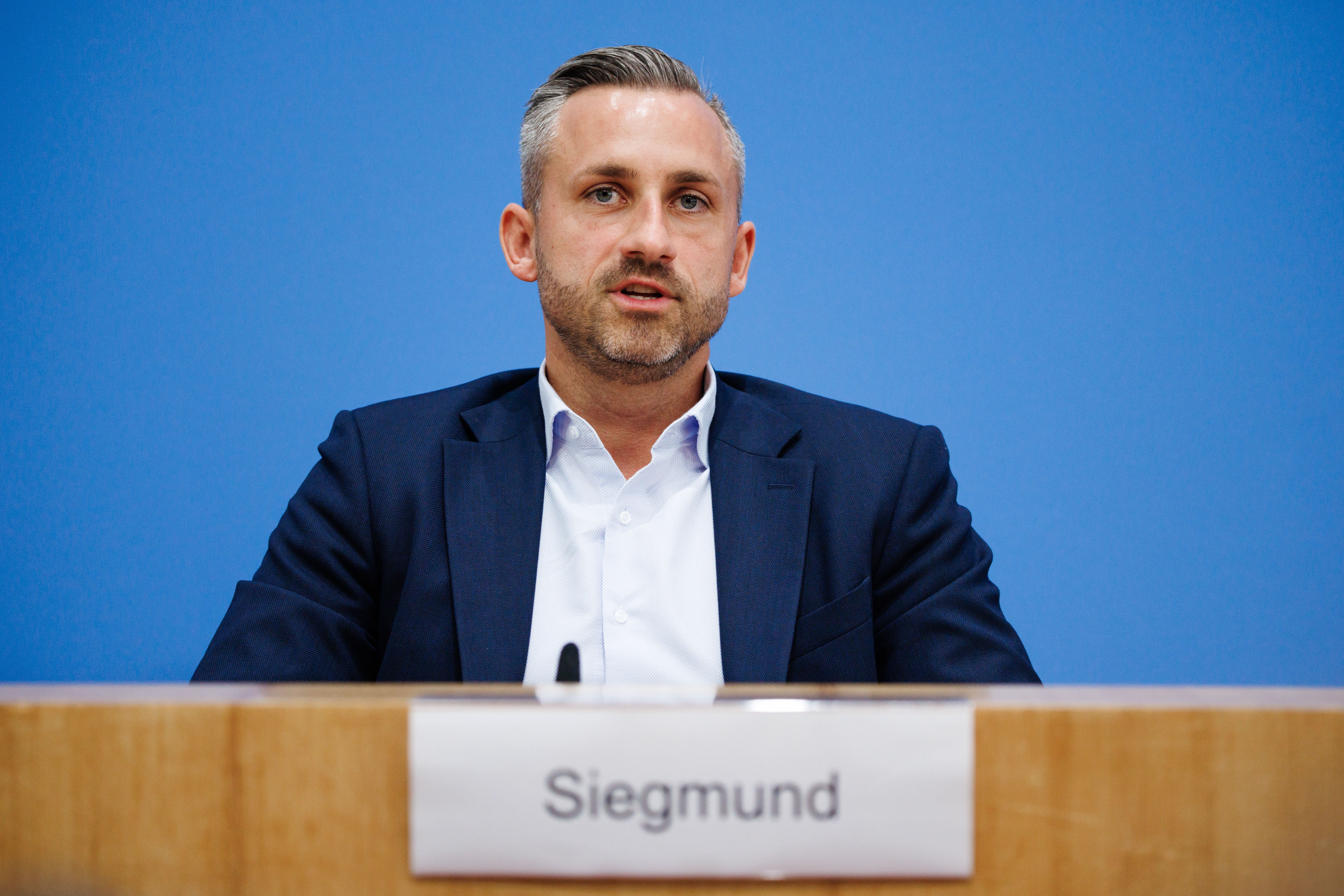 Ein Mann im Anzug spricht auf einer Pressekonferenz, Namensschild "Siegmund" vor ihm.