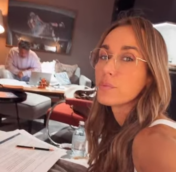 Eine Frau mit Brille sitzt am Tisch, im Hintergrund arbeitet ein Mann an einem Laptop auf dem Sofa.
