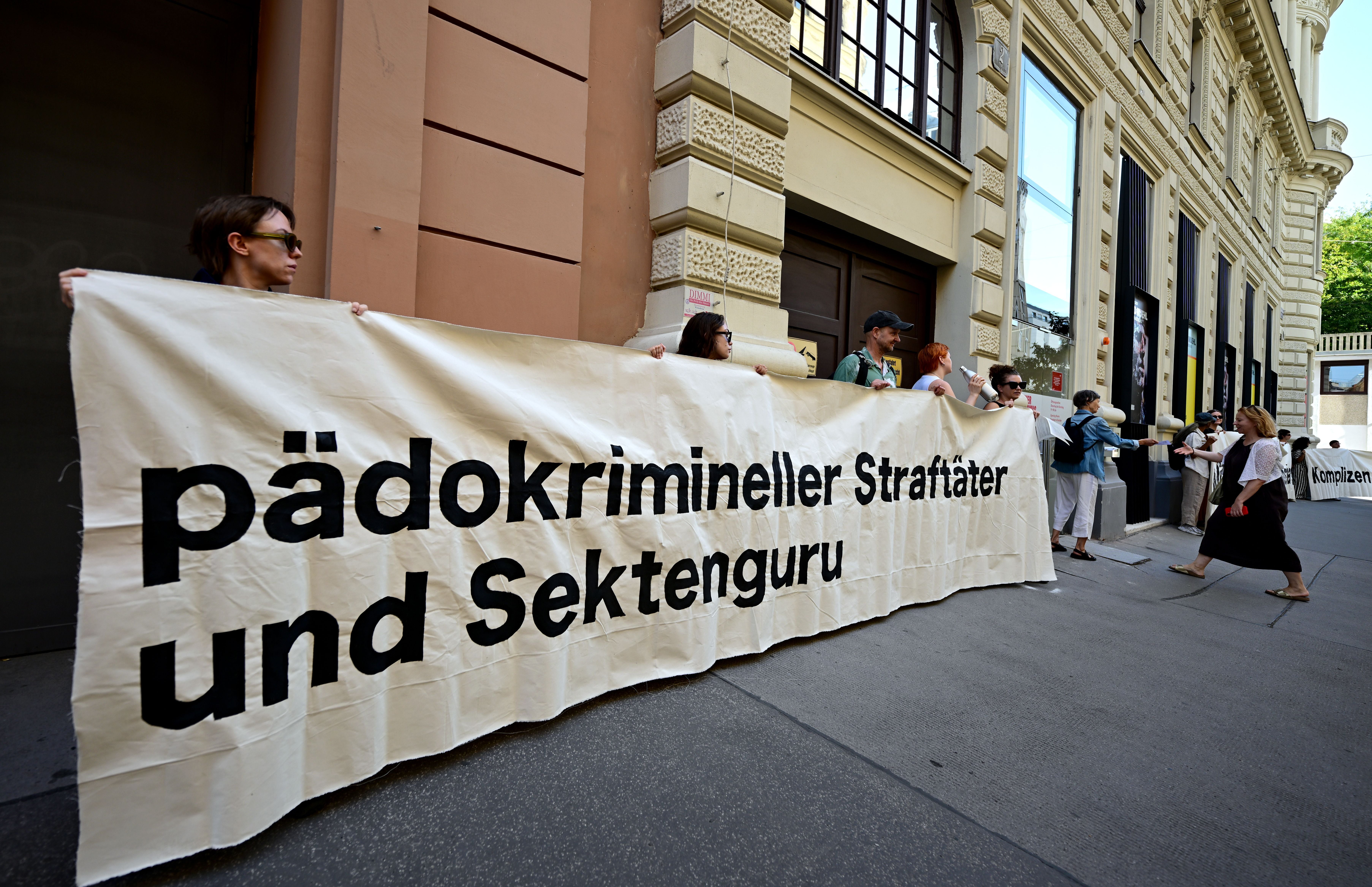 Mehrere Menschen halten bei einer Kundgebung ein großes Banner mit kritischer Aufschrift.