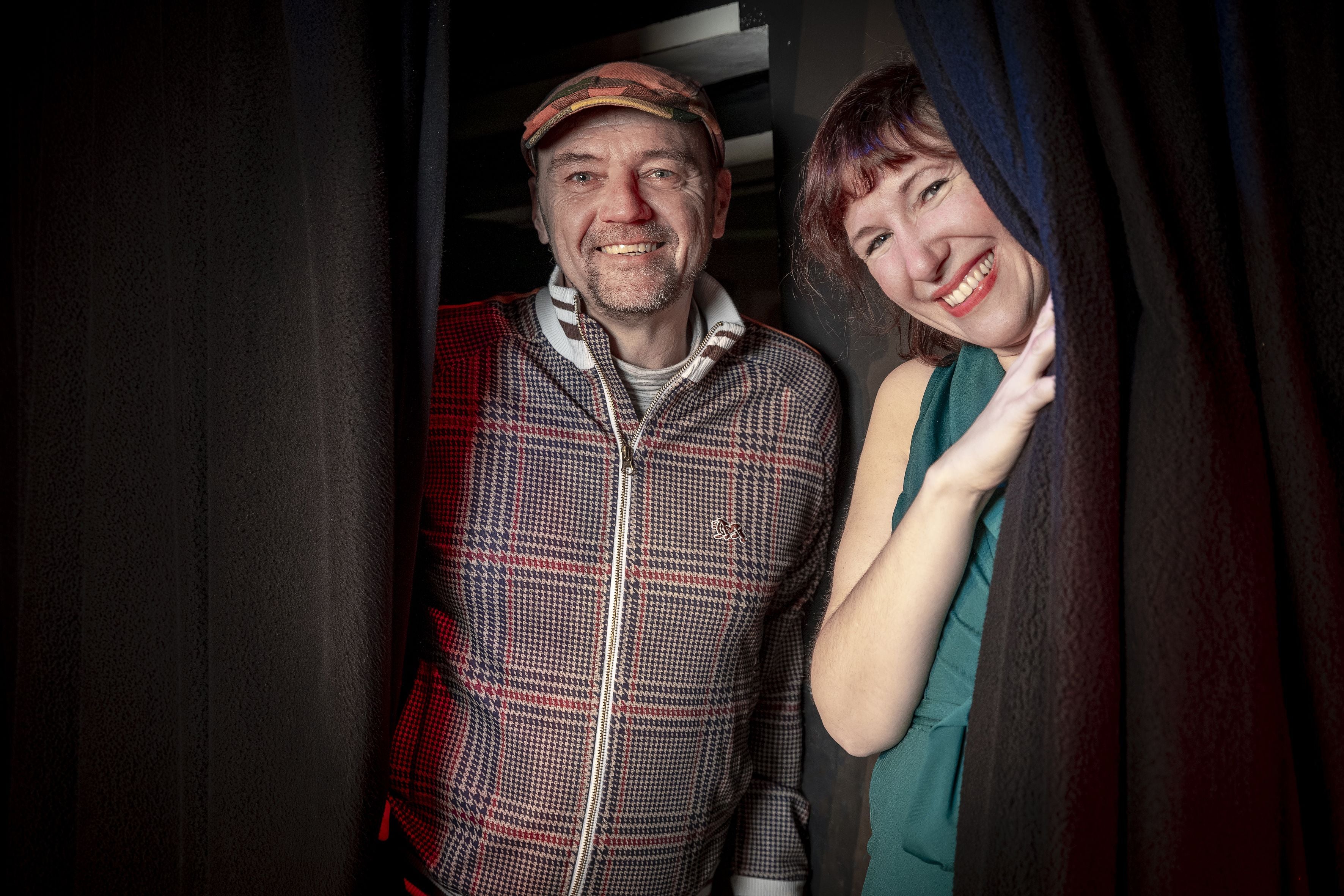 Ein Mann und eine Frau lächeln zwischen schwarzen Vorhängen in die Kamera.