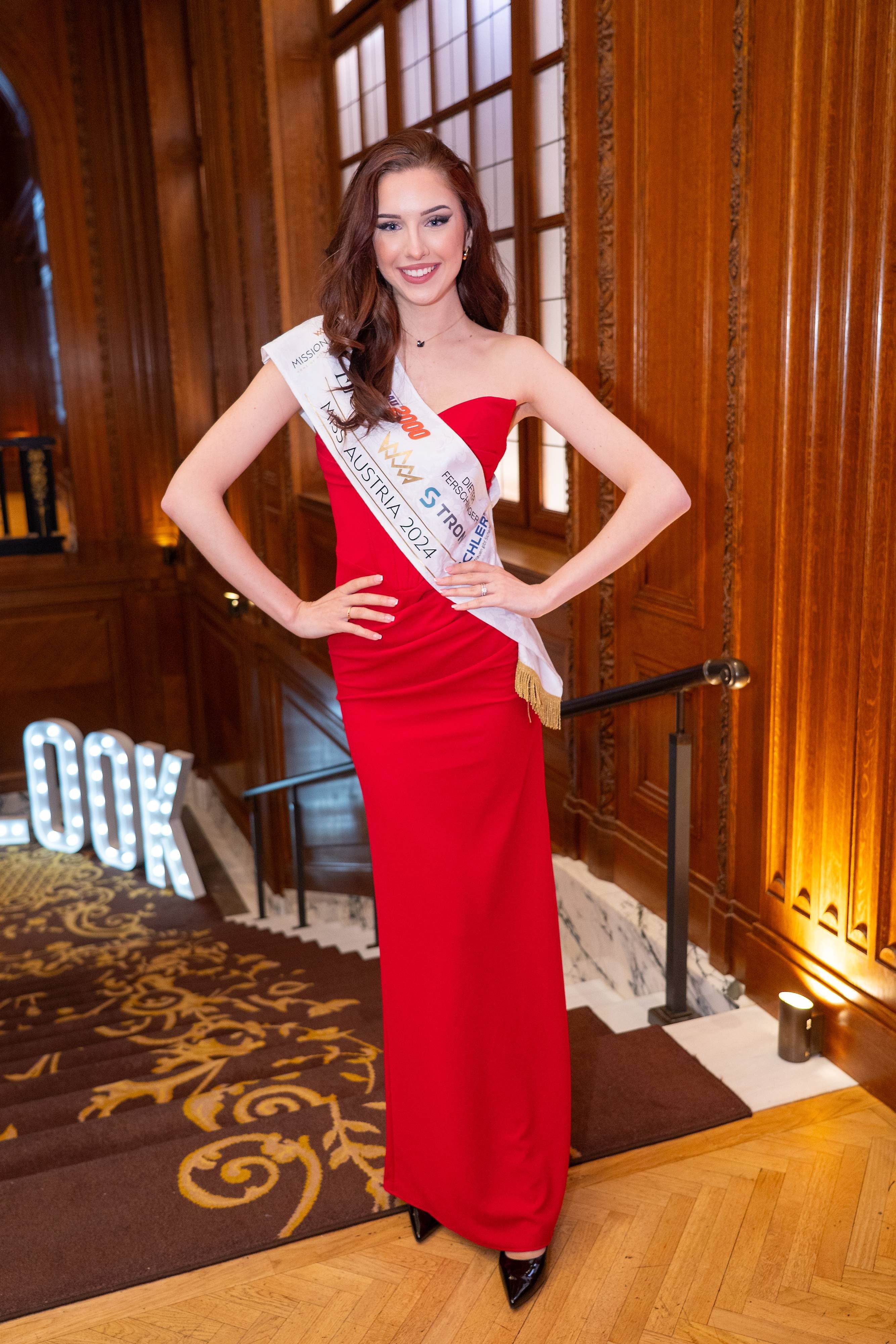 Junge Frau in rotem Kleid mit Schärpe „Miss Austria 2024“ posiert bei einer Preisverleihung.