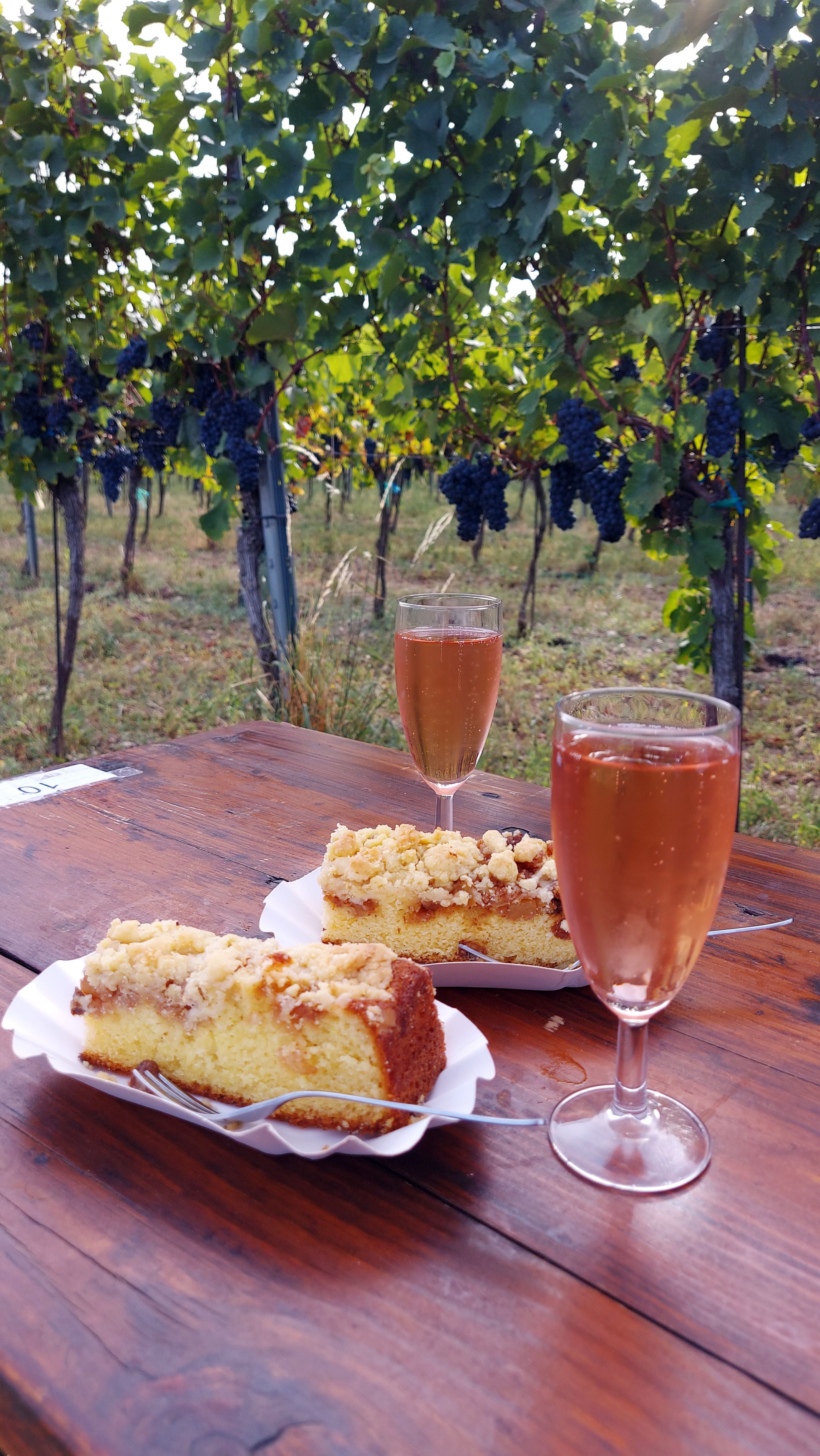 Zwei Gläser Sekt und Streuselkuchen auf Holztisch im Weingarten mit Trauben im Hintergrund.
