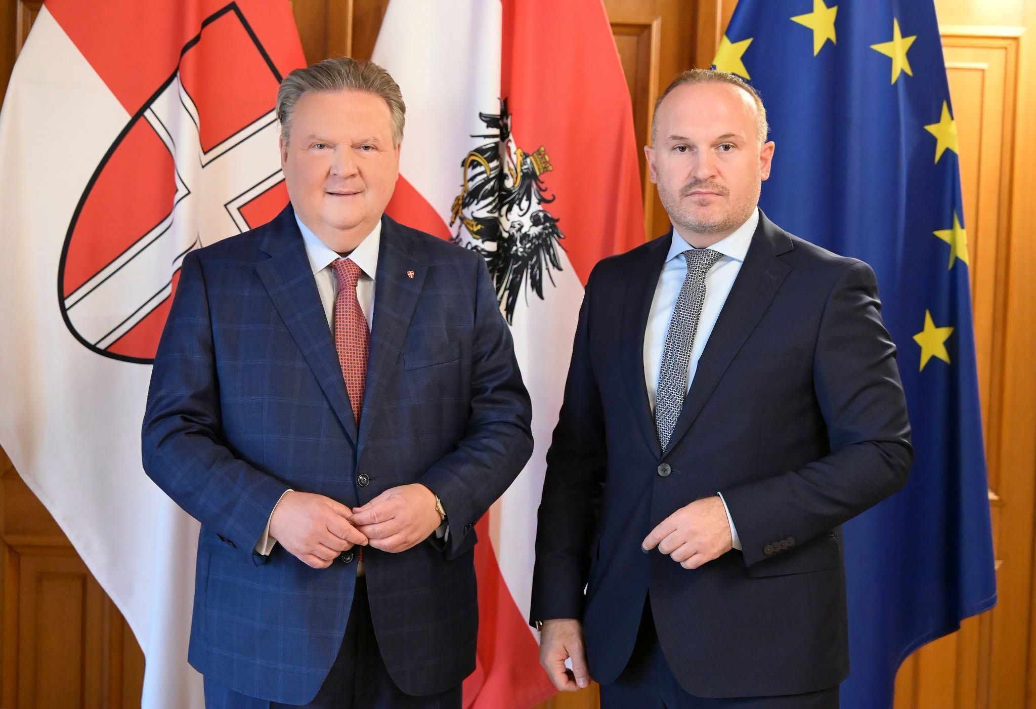 Zwei Männer im Anzug stehen vor österreichischen und EU-Flaggen und blicken in die Kamera.