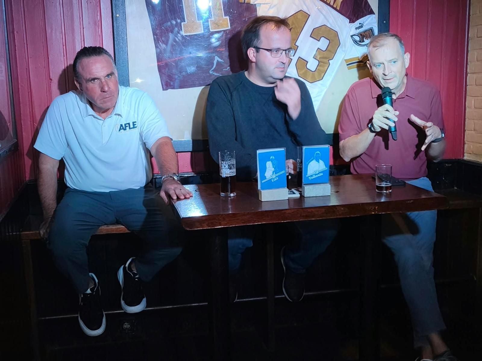 Drei Männer sitzen an einem Tisch, einer spricht mit Mikrofon beim Fan-Talk in gemütlicher Atmosphäre.