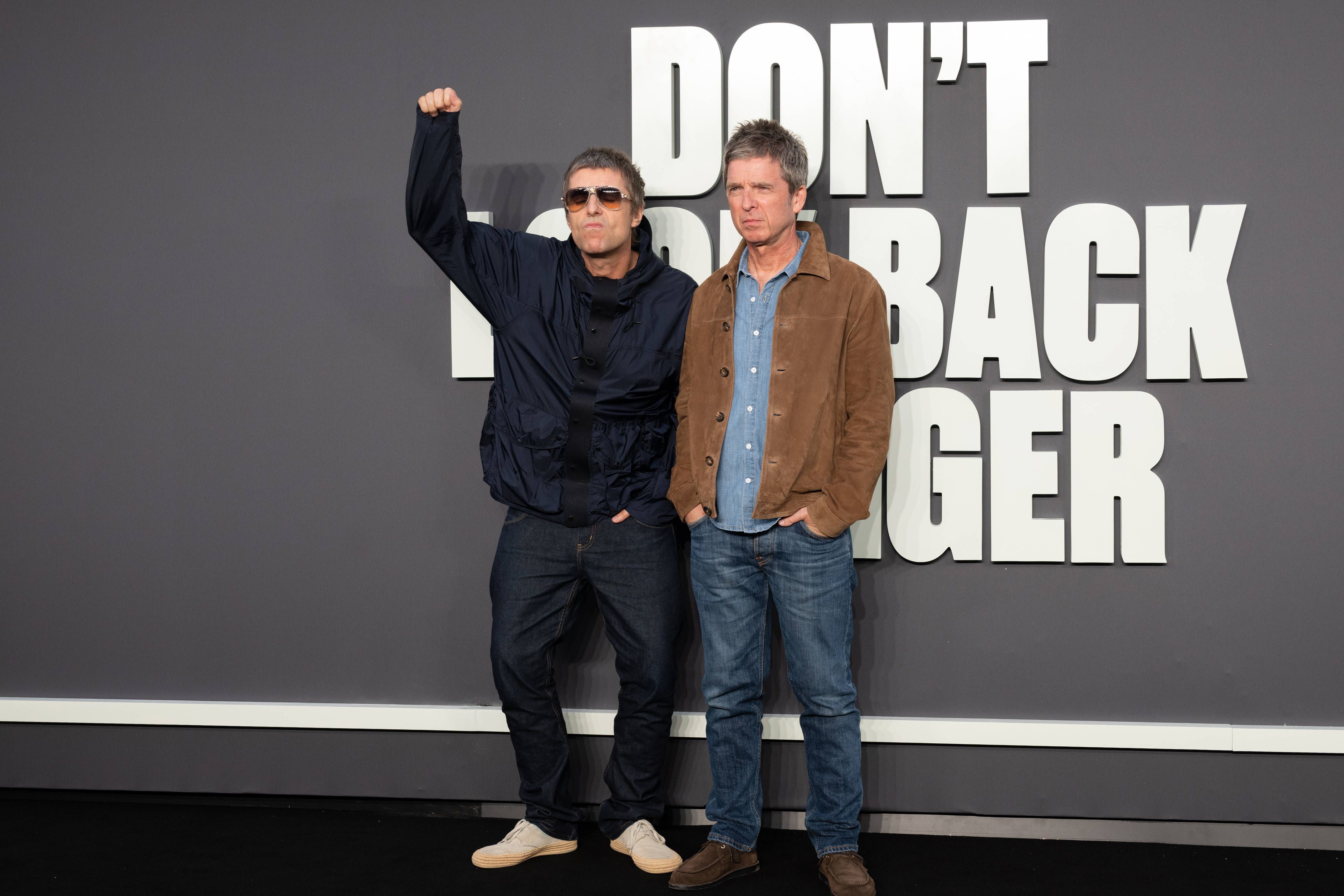 Zwei Männer posieren vor einer Wand mit der Aufschrift „DON’T LOOK BACK IN ANGER“.