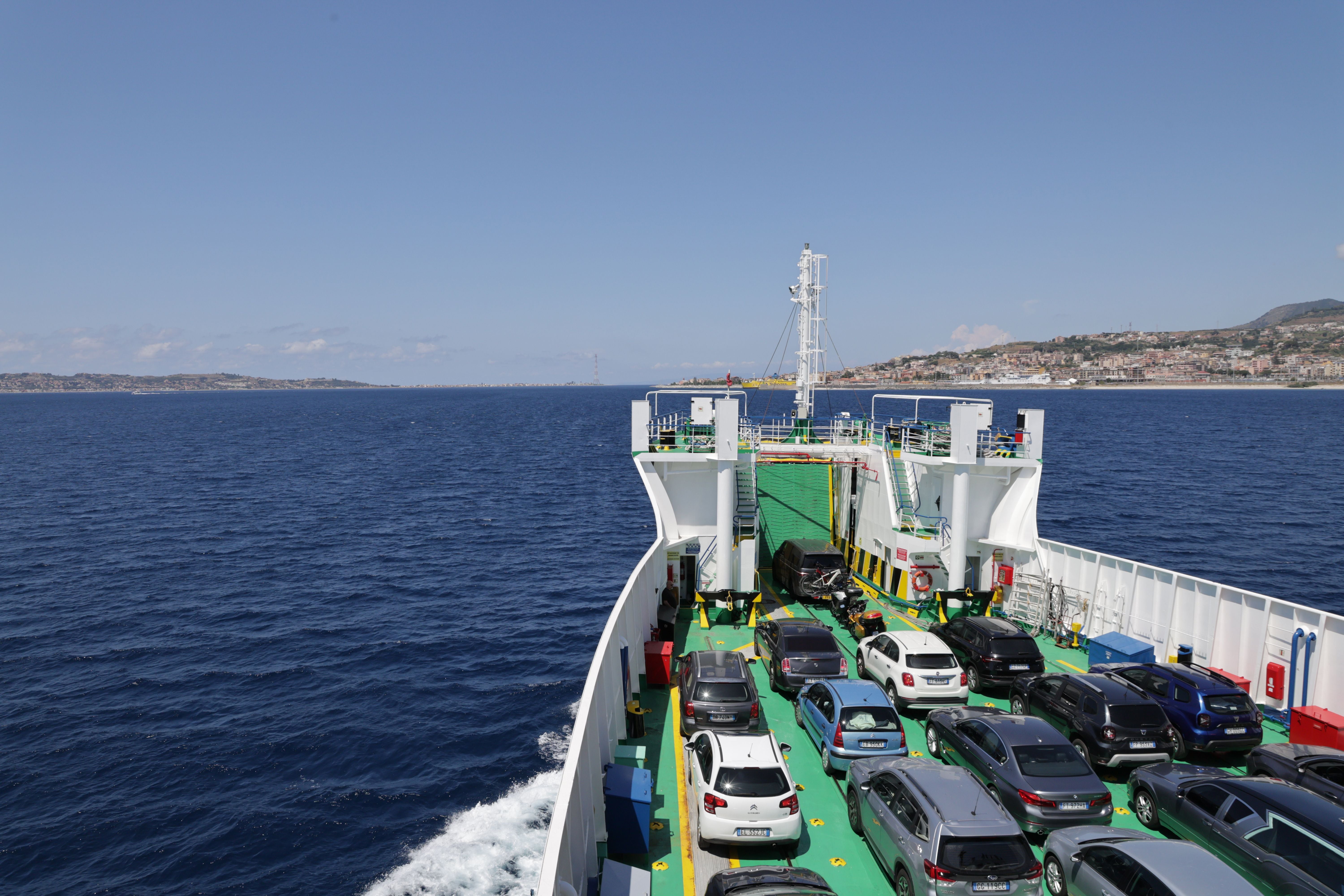 Autos stehen auf einer Fähre über das Meer, im Hintergrund ist Kalabrien an der Küste zu sehen.
