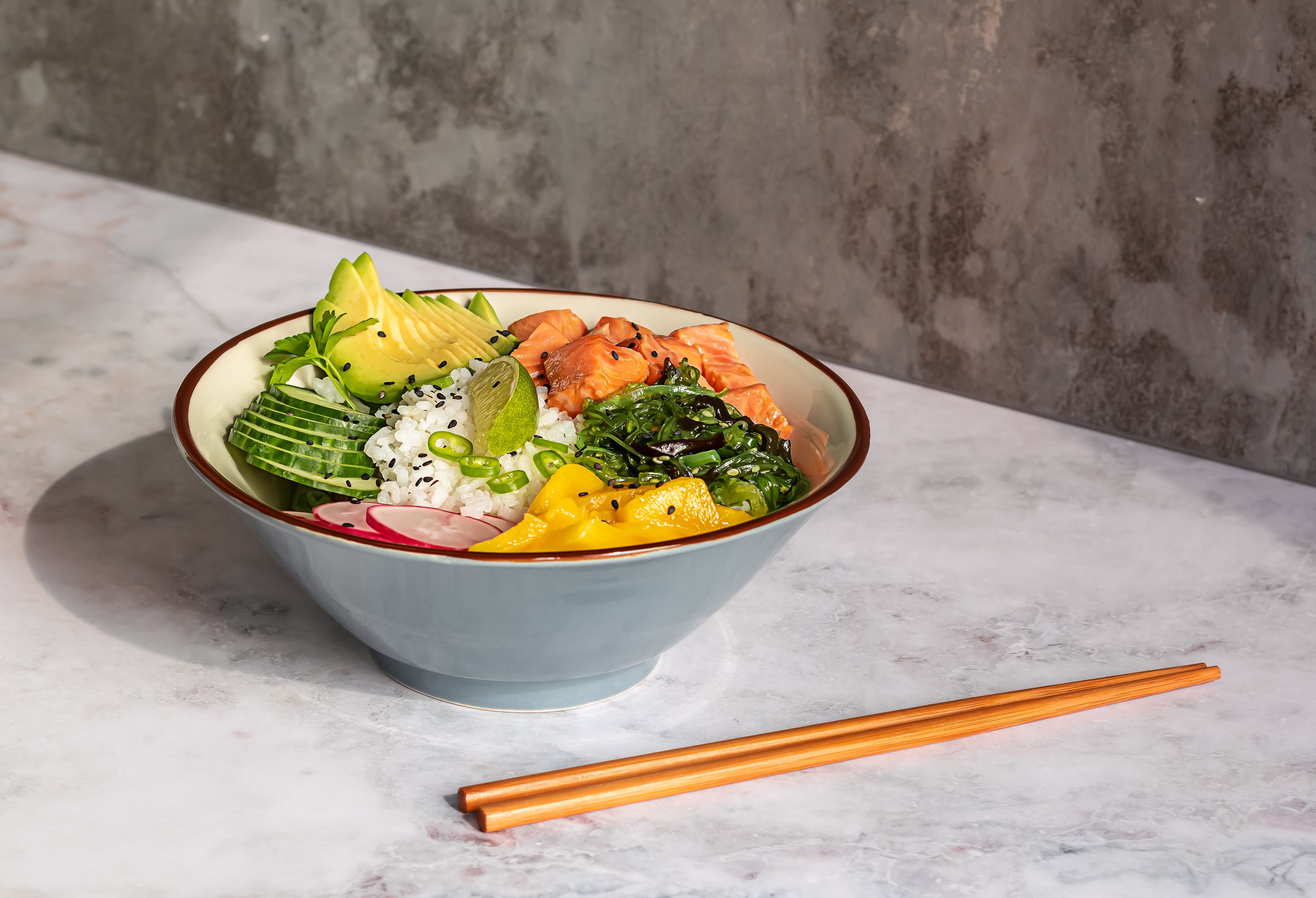 Poke Bowl mit Lachs, Reis, Avocado, Gurke, Mango, Algen und Radieschen auf Tisch mit Stäbchen.