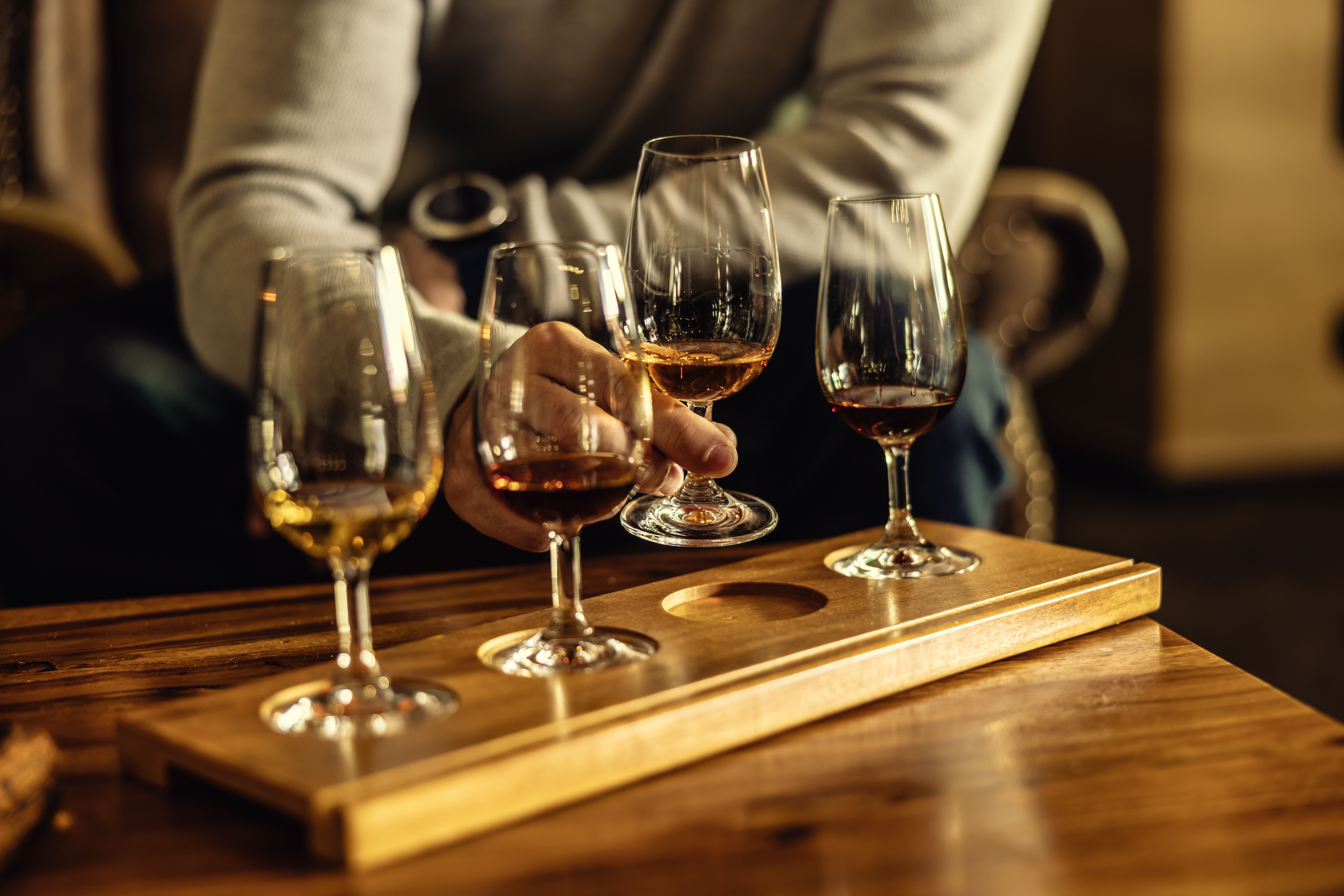 Eine Hand wählt ein Glas Rum aus einem Tasting-Set mit mehreren Gläsern auf Holztablett.